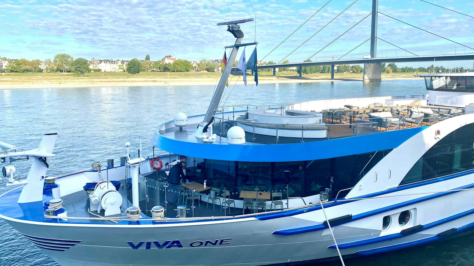 Ein Kreuzfahrtschiff liegt vertäut am Rheinufer, im Hintergrund eine gewaltige Autobahnbrücke.