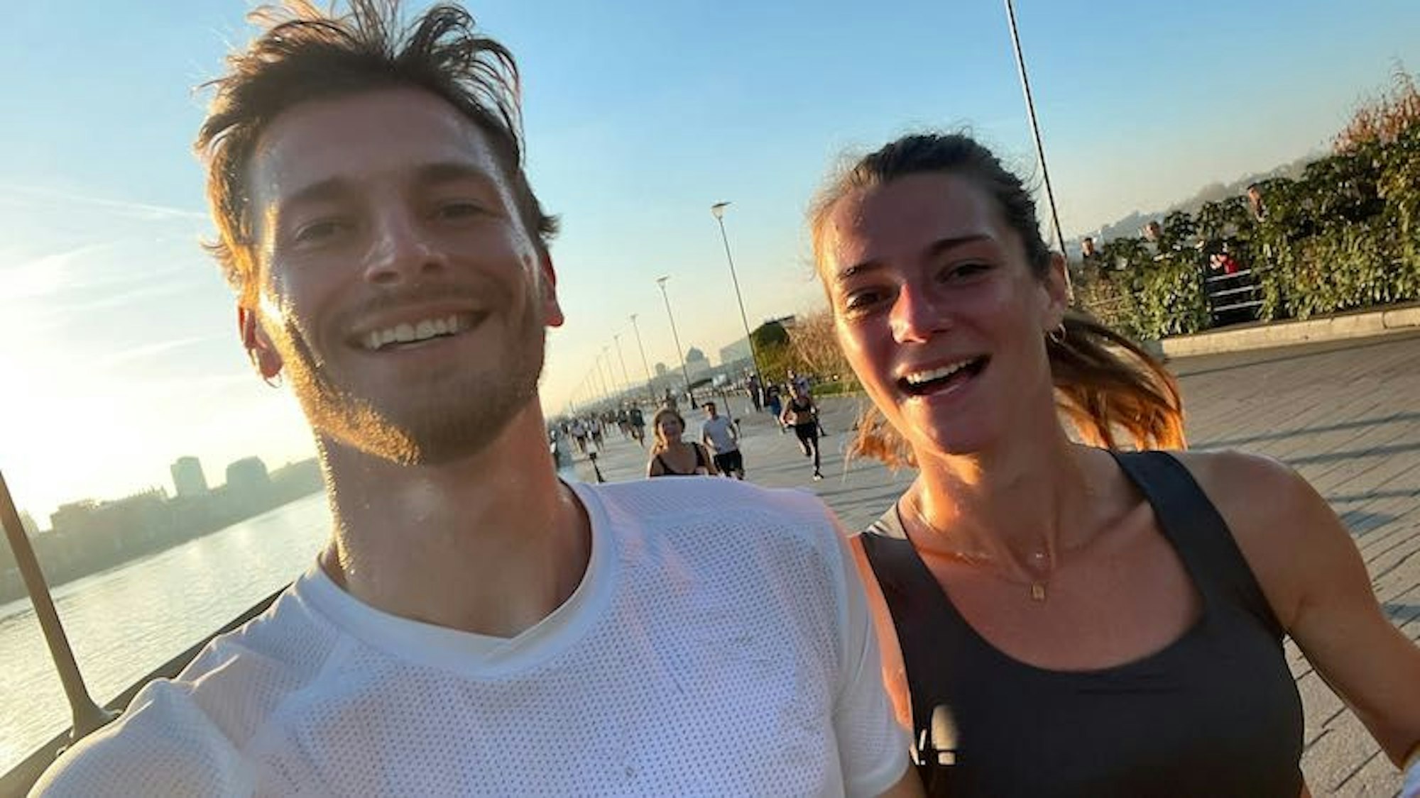 Ein junger Mann und eine junge Frau joggen gemeinsam an einem Flussufer entlang und schauen dabei fröhlich in die Kamera.