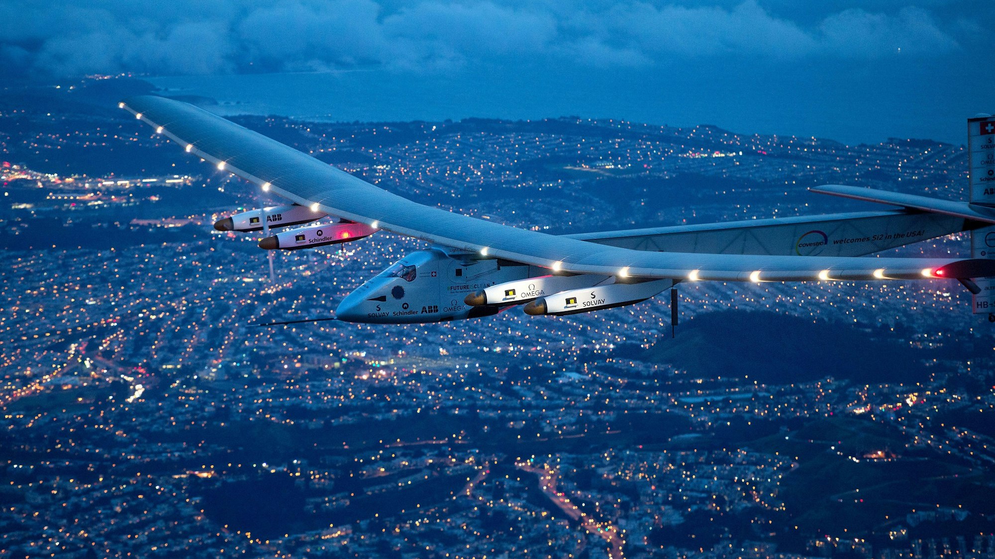 Ein beleuchtetes Flugzeug mit weiten Tragflächen gleitet bei Nacht über eine hellerleuchtete Stadt.