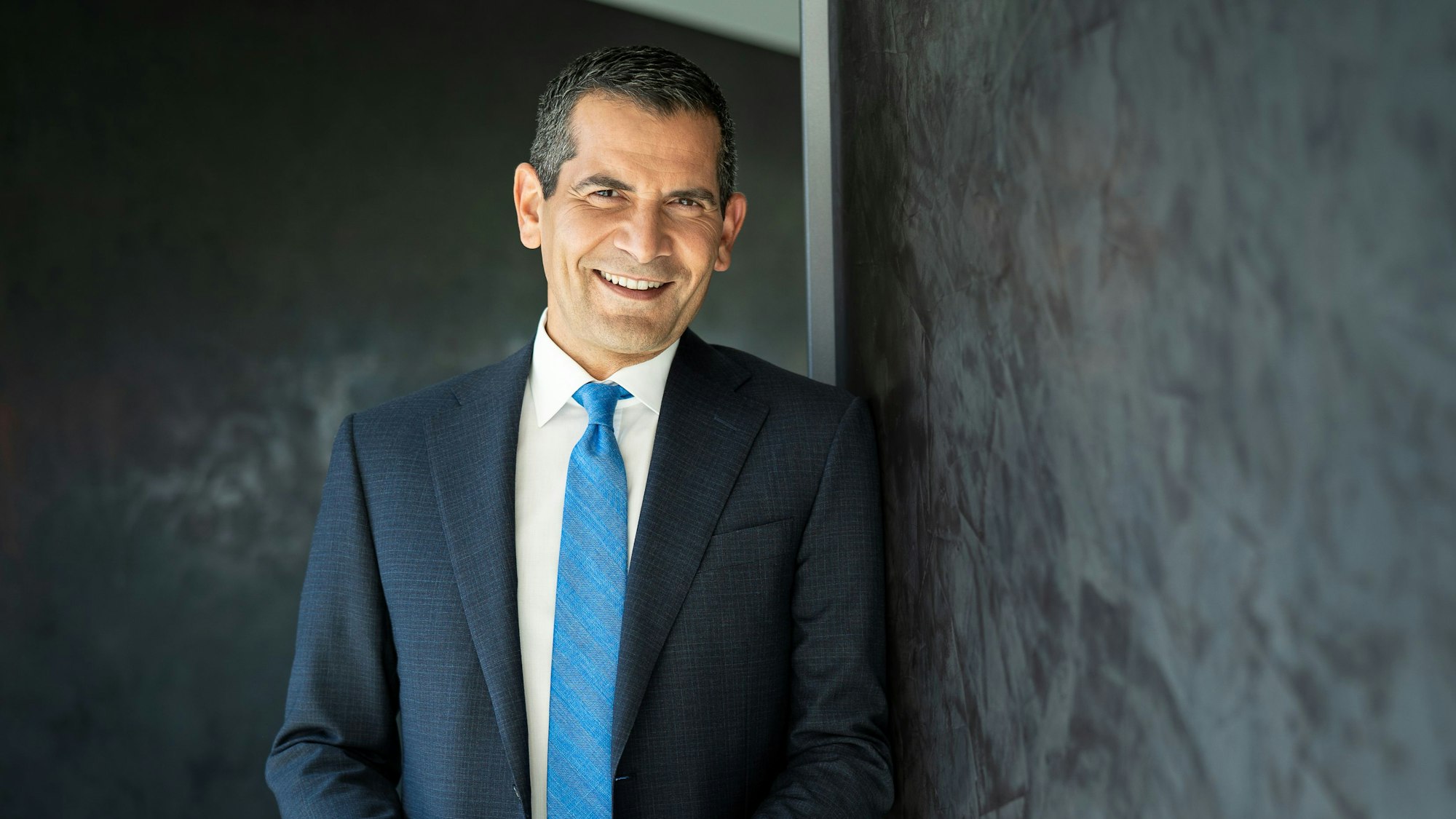 Ein Mann in dunkelblauem Anzug mit hellblauer Krawatte steht vor einer schiefergrauen Wand