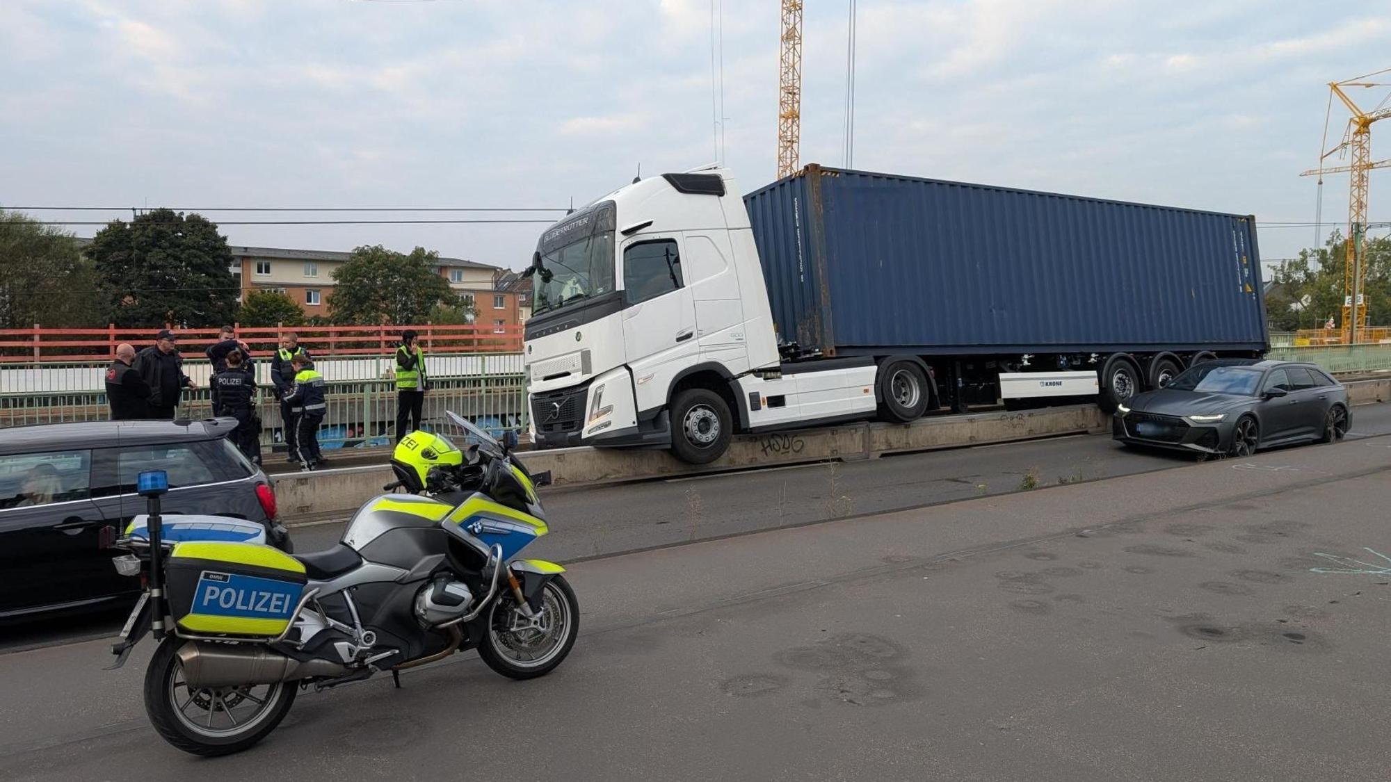 Ein Lkw steht mit zum Teil in der Luft hängenden Rädern auf einer niedrigen Betonwand. Einsatzkräfte der Polizei gucken sich das an.