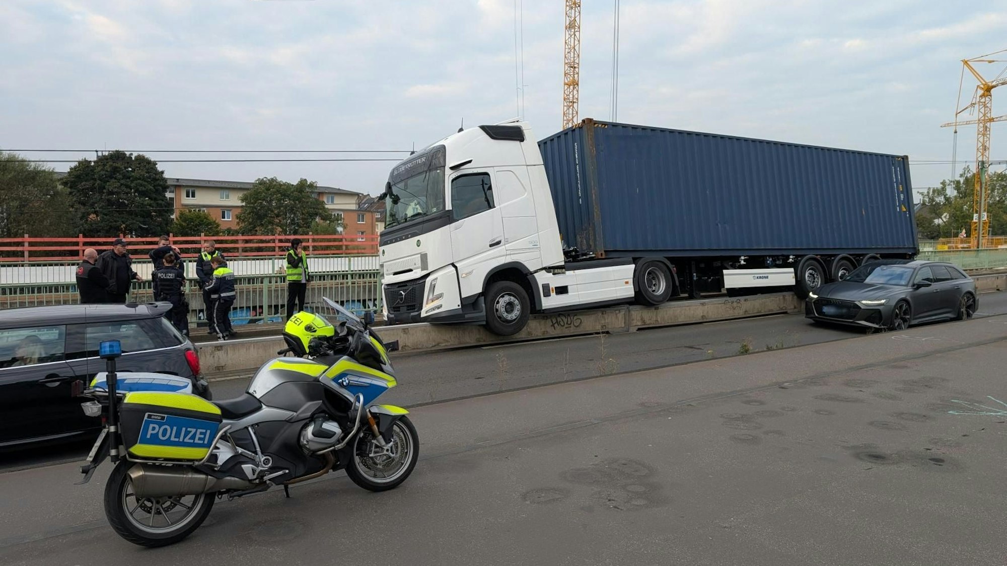 Ein Lkw steht mit zum Teil in der Luft hängenden Rädern auf einer niedrigen Betonwand. Einsatzkräfte der Polizei gucken sich das an.