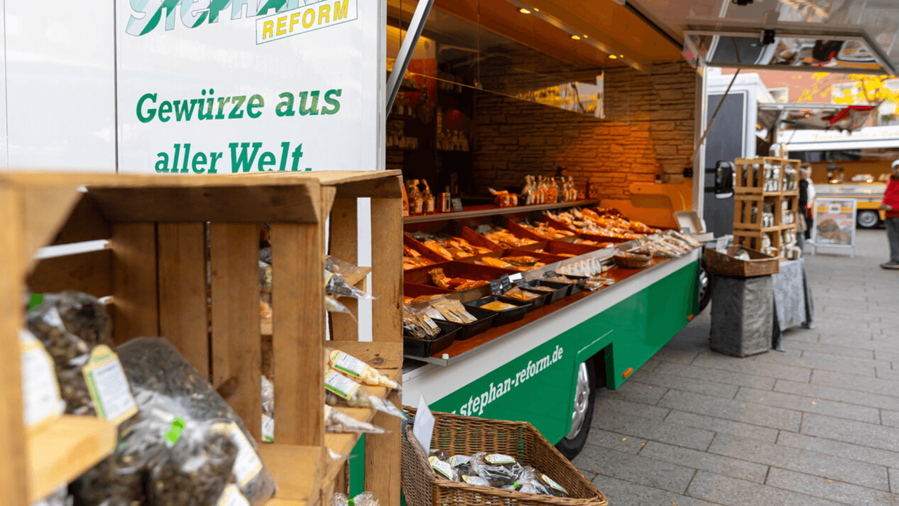 Auf dem Bild ist ein Marktstand zu sehen, der mit dem Slogan „Gewürze aus aller Welt“ wirbt. In Holzkisten und Auslagen werden zahlreiche verpackte Gewürze präsentiert, die zum Stöbern und Kaufen einladen.