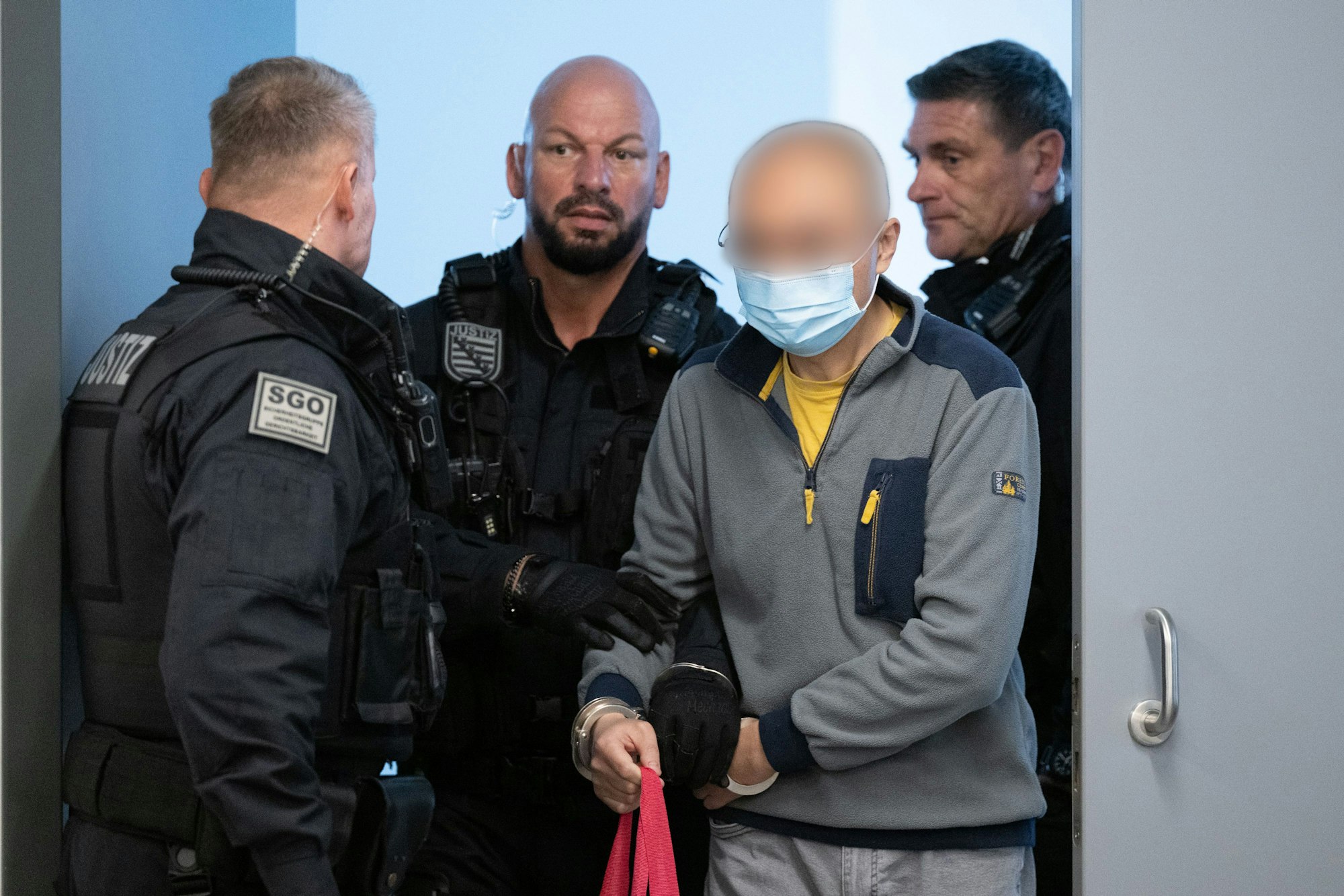 Drei Wachtmeister führen einen Mann in Handschellen in den Gerichtssaal.