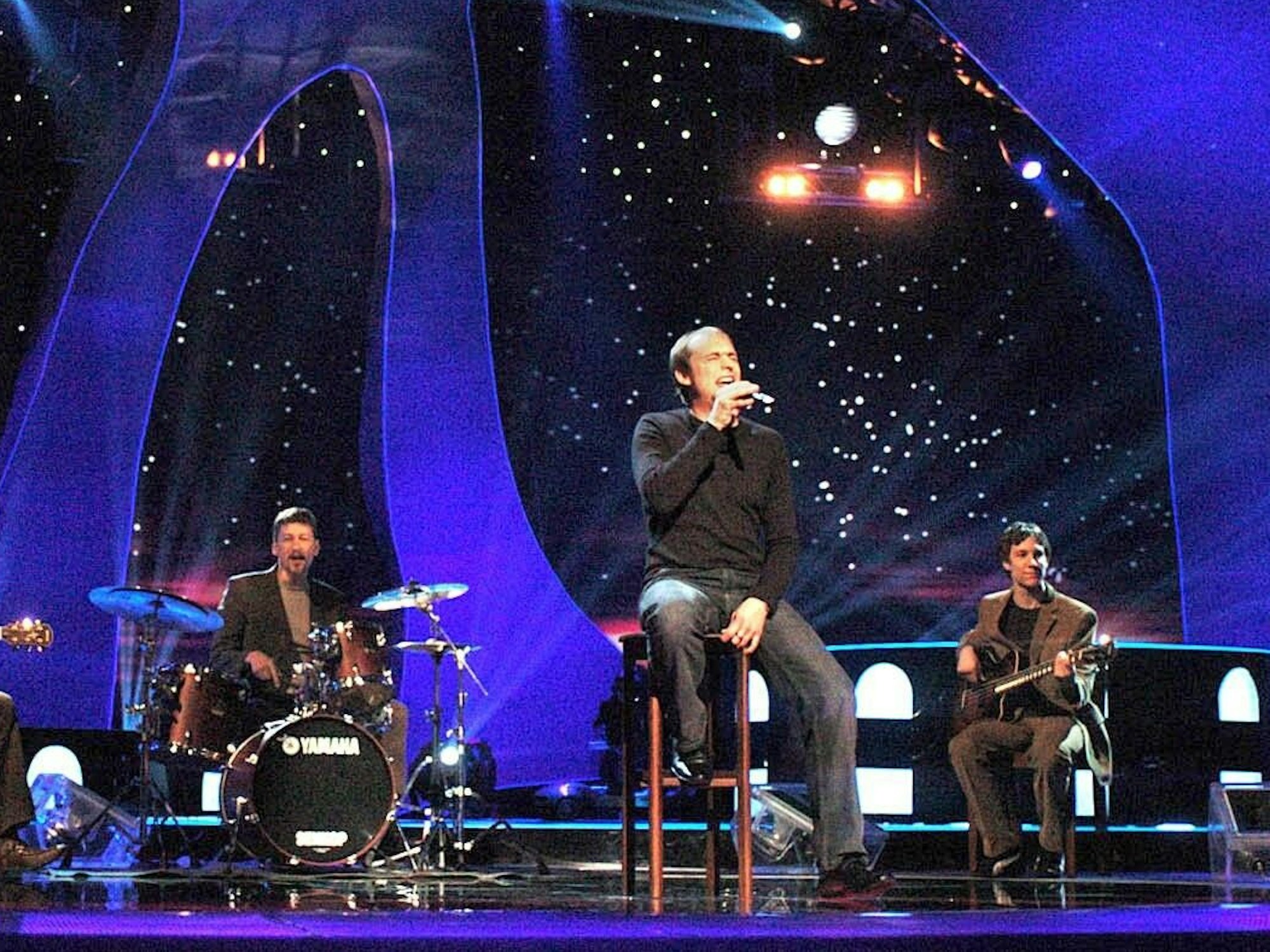 Ein Man sitzt auf einer Bühne auf einem Barhocker und singt von Musikern begleitet - im Hintergrund eine Kulisse mit Sternen und Lichtern.