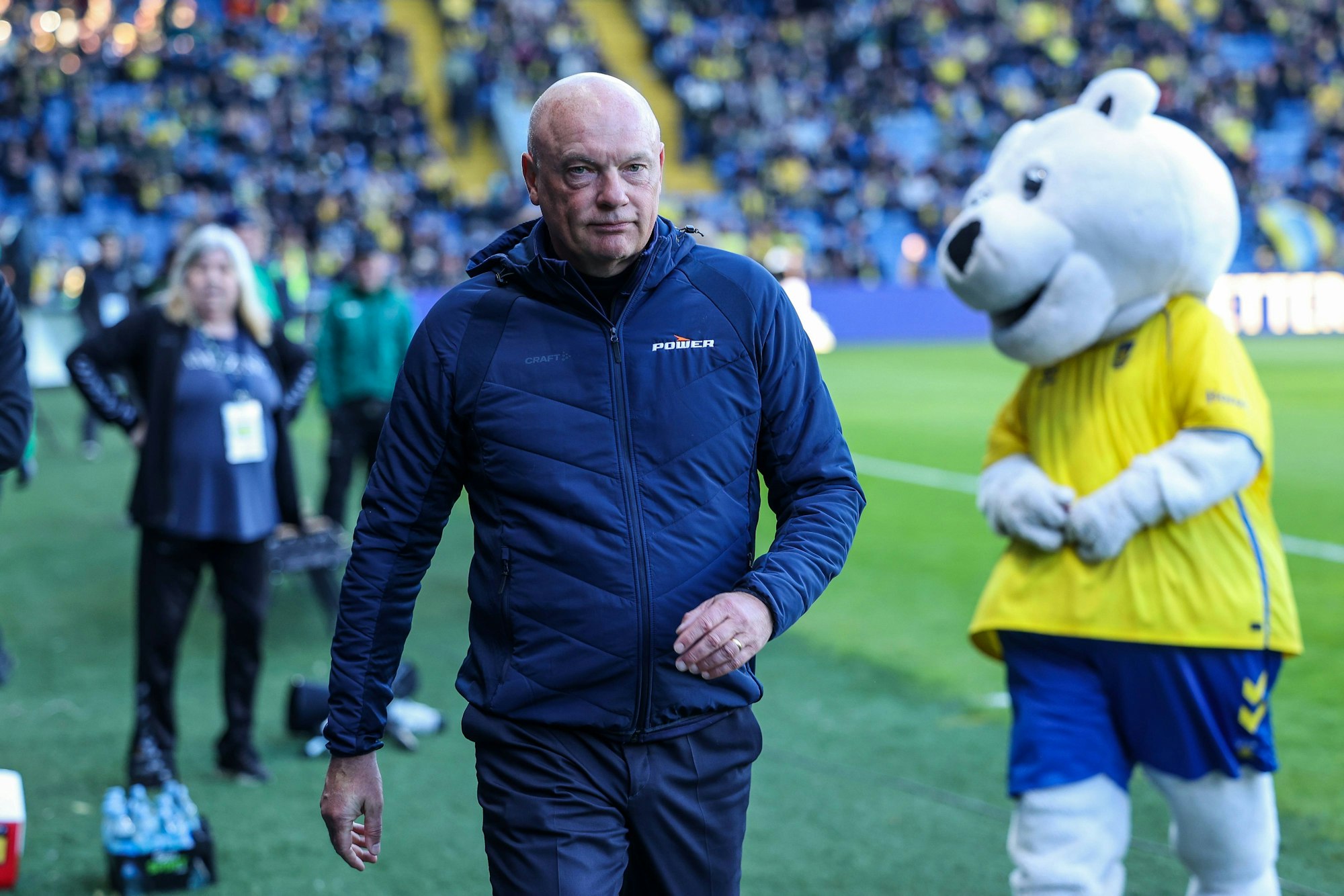 Uwe Rösler geht über den Platz, hinter ihm steht ein Maskottchen.