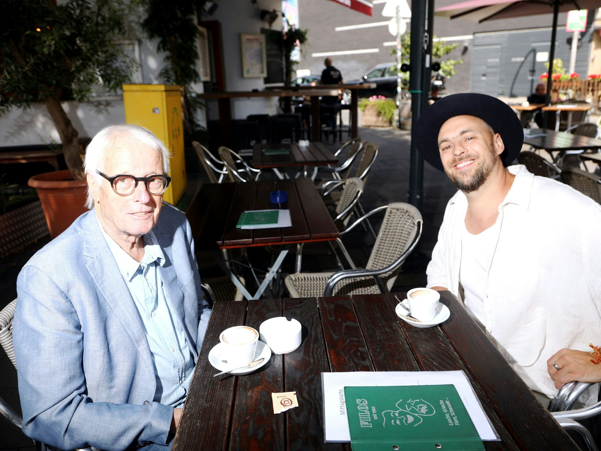Ein älterer Herr mit markanter schwarz umrandeter Brille im Jackett sitzt mit einem lachenden jungen Mann in weißem Hemd mit schwarzem Hut in einem Straßencafé.
