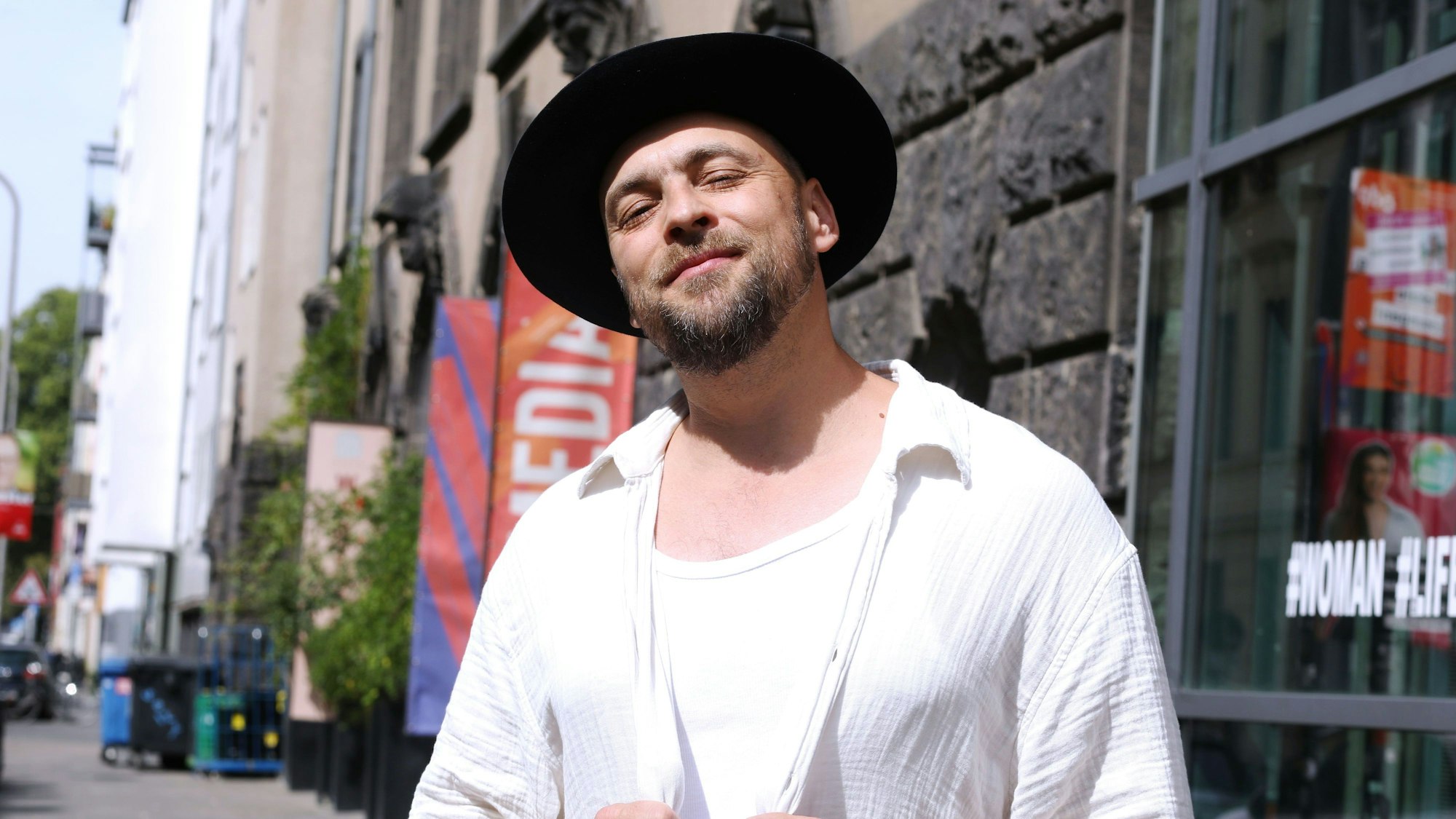 Ein bärtiger Mann mittleren Alters mit schwarzem Hut und offenem Hemd steht vor einer Häuserzeile und guckt in die Kamera.