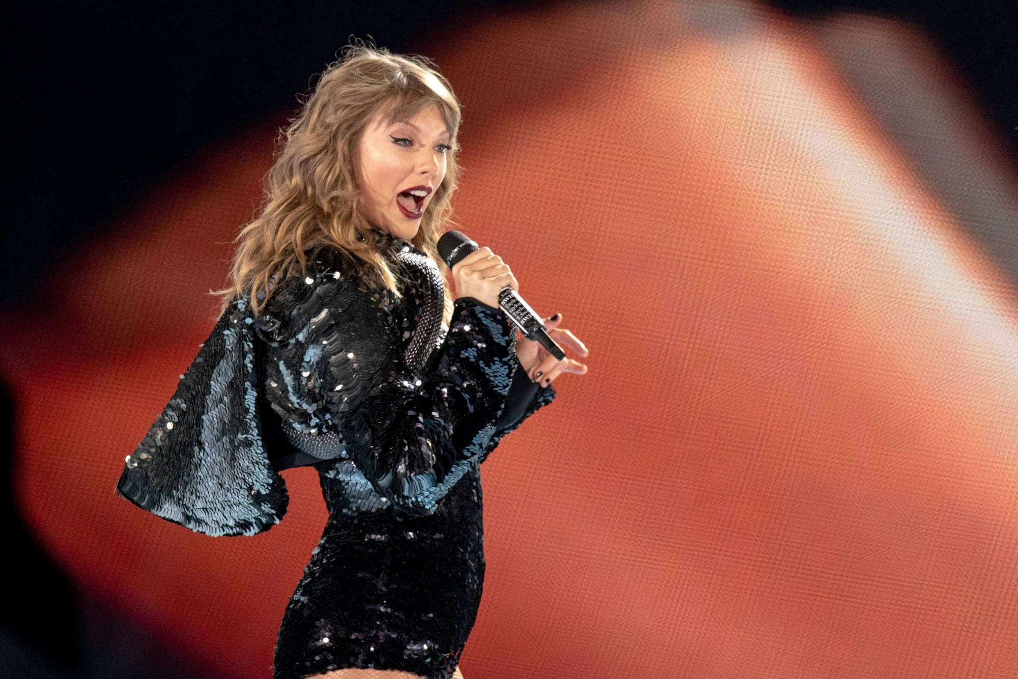 Taylor Swift singt leidenschaftlich im schwarzen Paillettenoutfit vor orangefarbenem Hintergrund