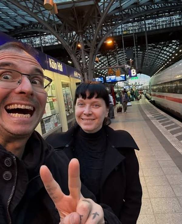 Ein Mann zeigt mit zwei Fingern ein V, dahinter steht eine Frau. Beide sind am Kölner Hauptbahnhof.