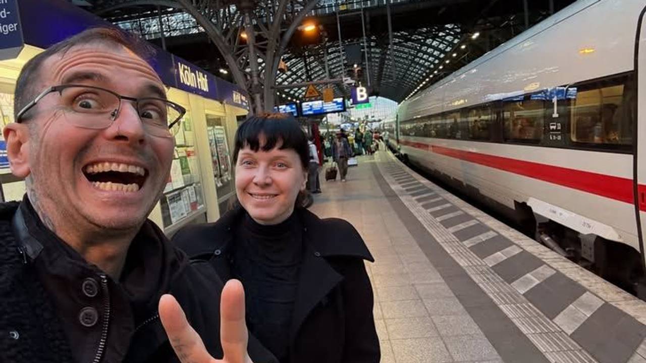 Ein Mann zeigt mit zwei Fingern ein V, dahinter steht eine Frau. Beide sind am Kölner Hauptbahnhof.