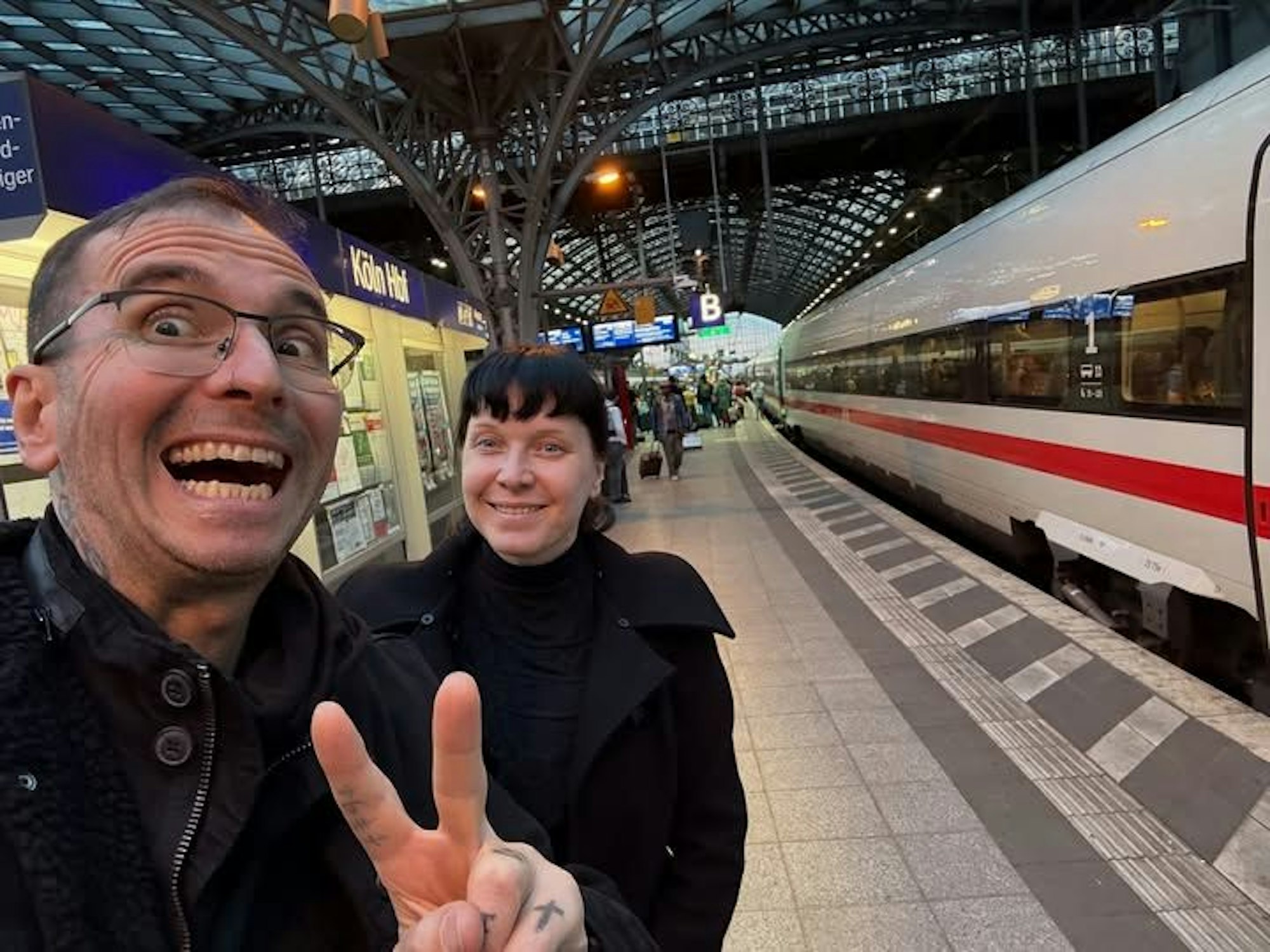 Ein Mann zeigt mit zwei Fingern ein V, dahinter steht eine Frau. Beide sind am Kölner Hauptbahnhof.