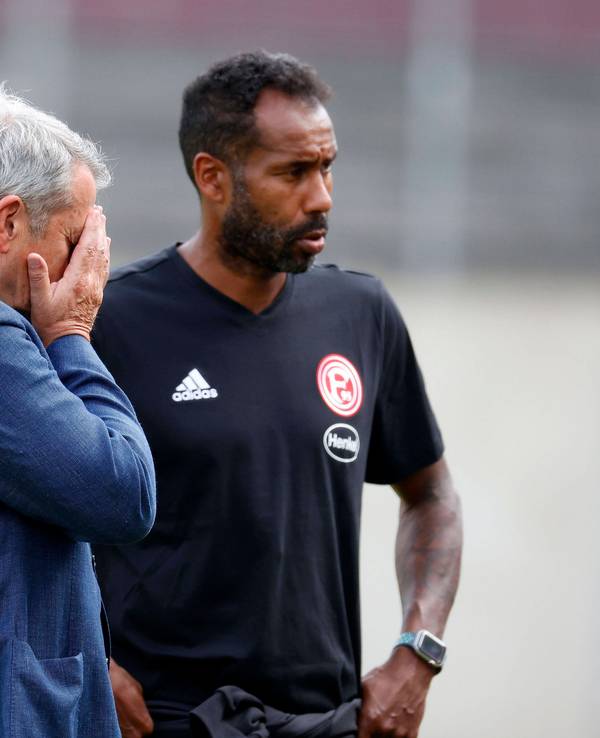 Daniel Thioune steht auf dem Trainingsplatz von Fortuna Düsseldorf neben F95-Boss Klaus Allofs, der die Hände vor das Gesicht schlägt.
