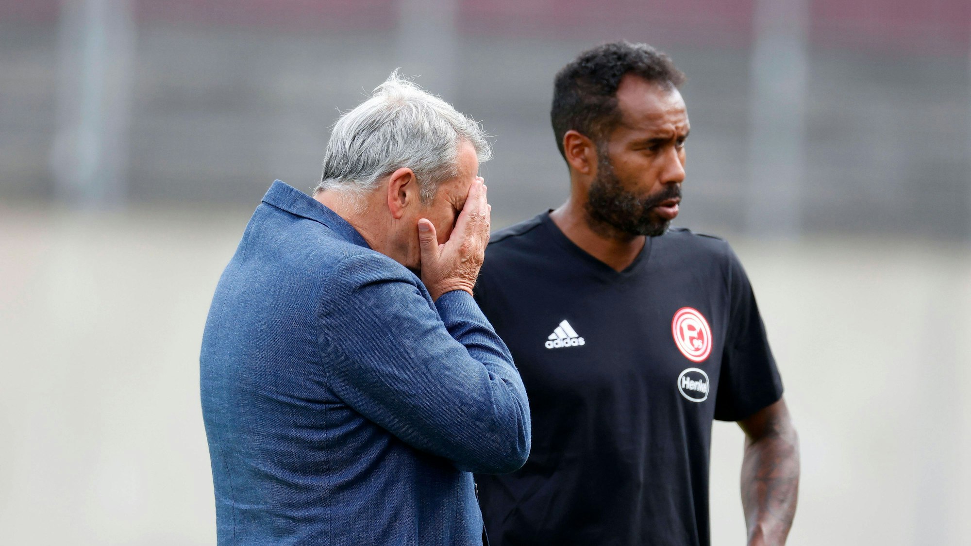 Daniel Thioune steht auf dem Trainingsplatz von Fortuna Düsseldorf neben F95-Boss Klaus Allofs, der die Hände vor das Gesicht schlägt.