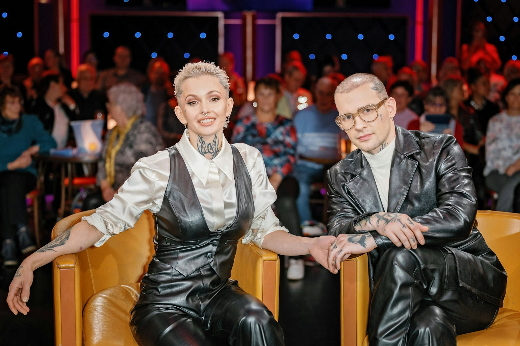 Eric Stehfest und Frau mit Tattoos in Lederoutfits lächeln in gelben Sesseln