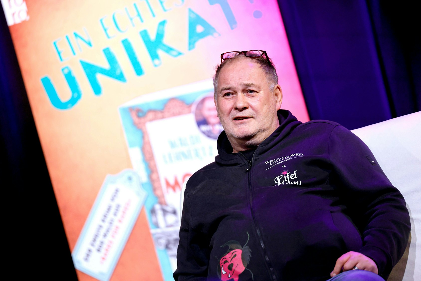 Walter Lehnertz sitzt in schwarzem Hoodie vor einem Plakat mit „UNIKAT“-Schriftzug