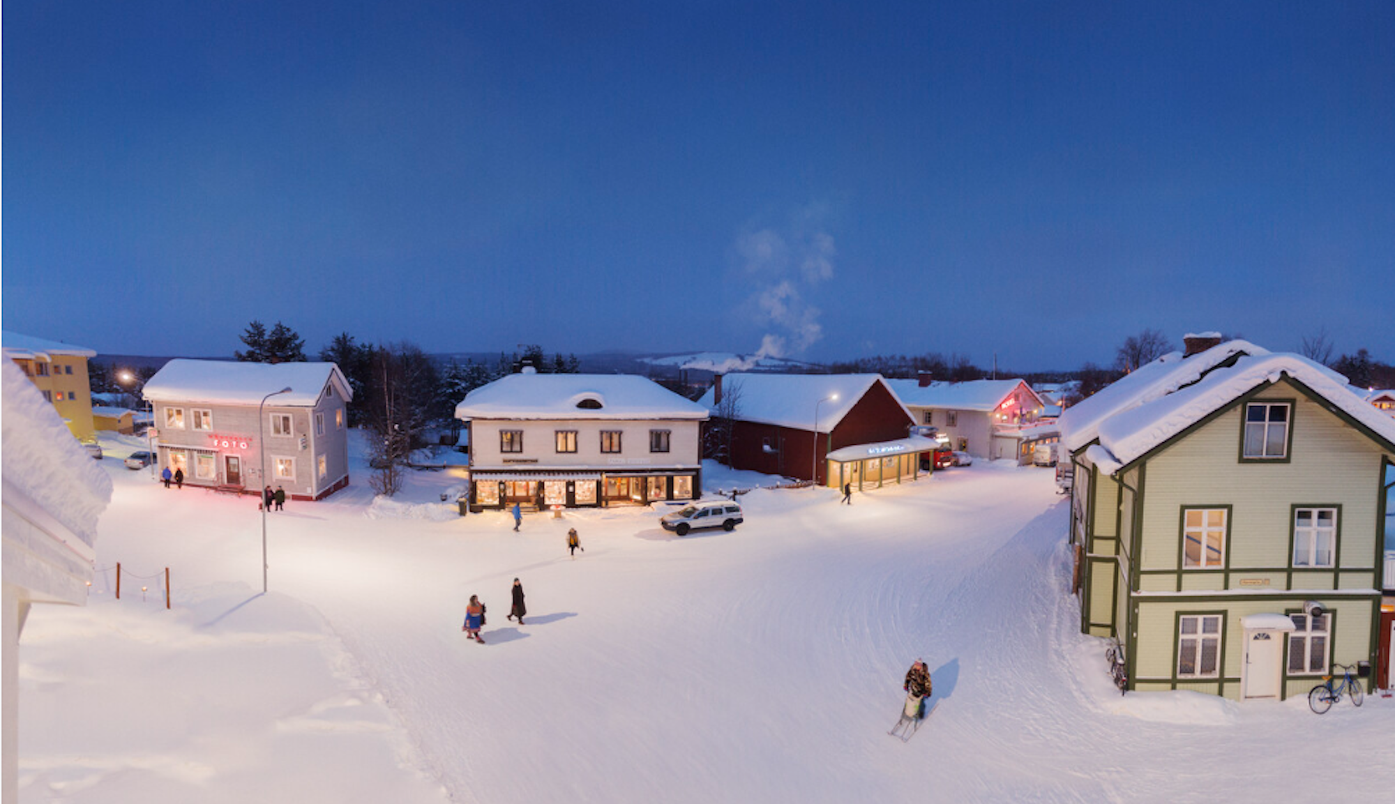 Das Bild zeigt ein verschneites Dorf in der Dämmerung. Warme Lichter erhellen die gemütlichen Holzhäuser, während Menschen durch den tiefen Schnee spazieren.
