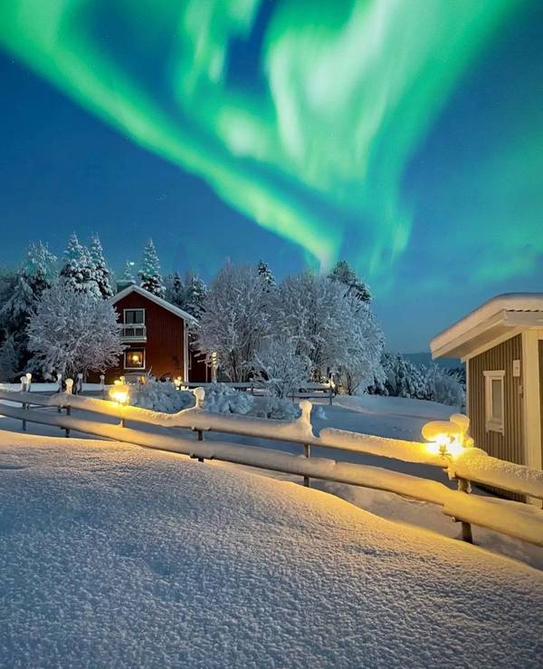 Das Bild zeigt eine Winterlandschaft. Eine schneebedeckte Straße führt an gemütlichen Holzhäusern vorbei. Über der Szene tanzen grüne Nordlichter am klaren Nachthimmel.