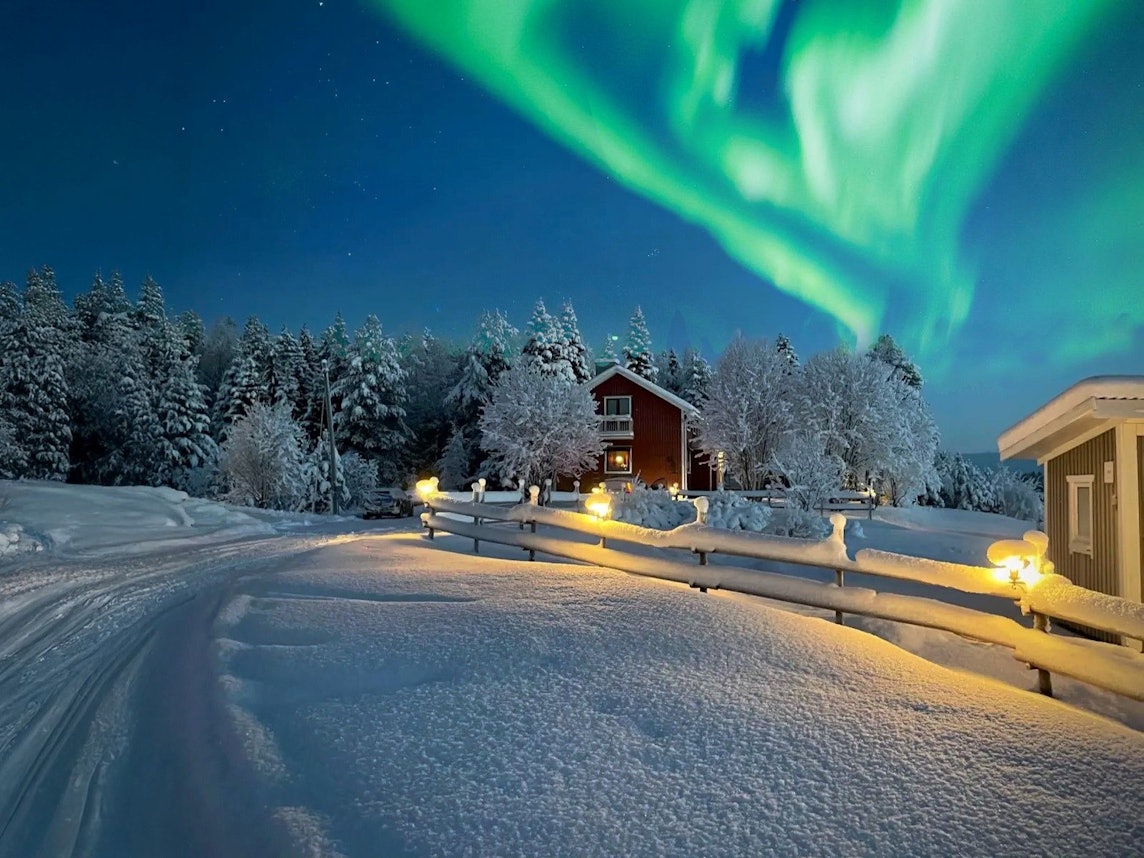 Das Bild zeigt eine Winterlandschaft. Eine schneebedeckte Straße führt an gemütlichen Holzhäusern vorbei. Über der Szene tanzen grüne Nordlichter am klaren Nachthimmel.