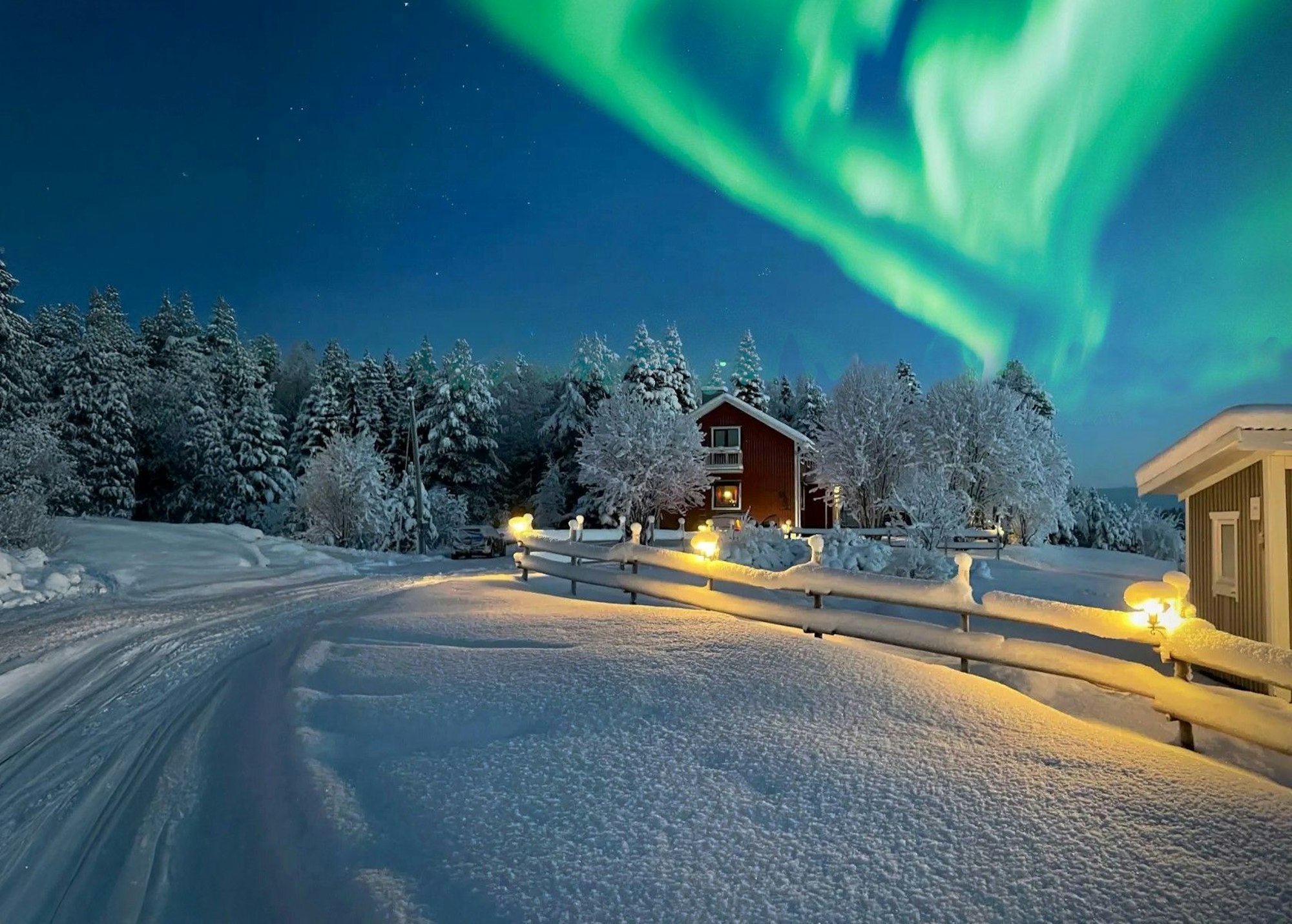 Das Bild zeigt eine Winterlandschaft. Eine schneebedeckte Straße führt an gemütlichen Holzhäusern vorbei. Über der Szene tanzen grüne Nordlichter am klaren Nachthimmel.
