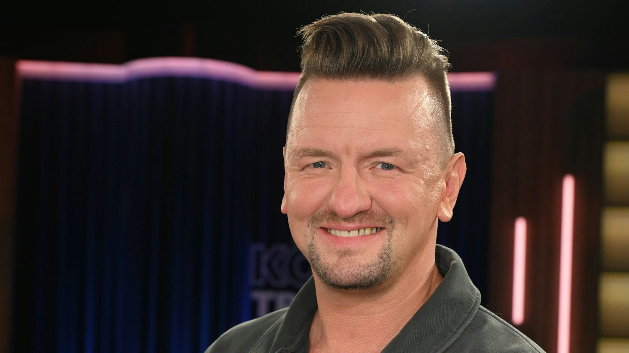 Ein Mann mit Haartolle und Dreitagebart lächelt in die Kamera