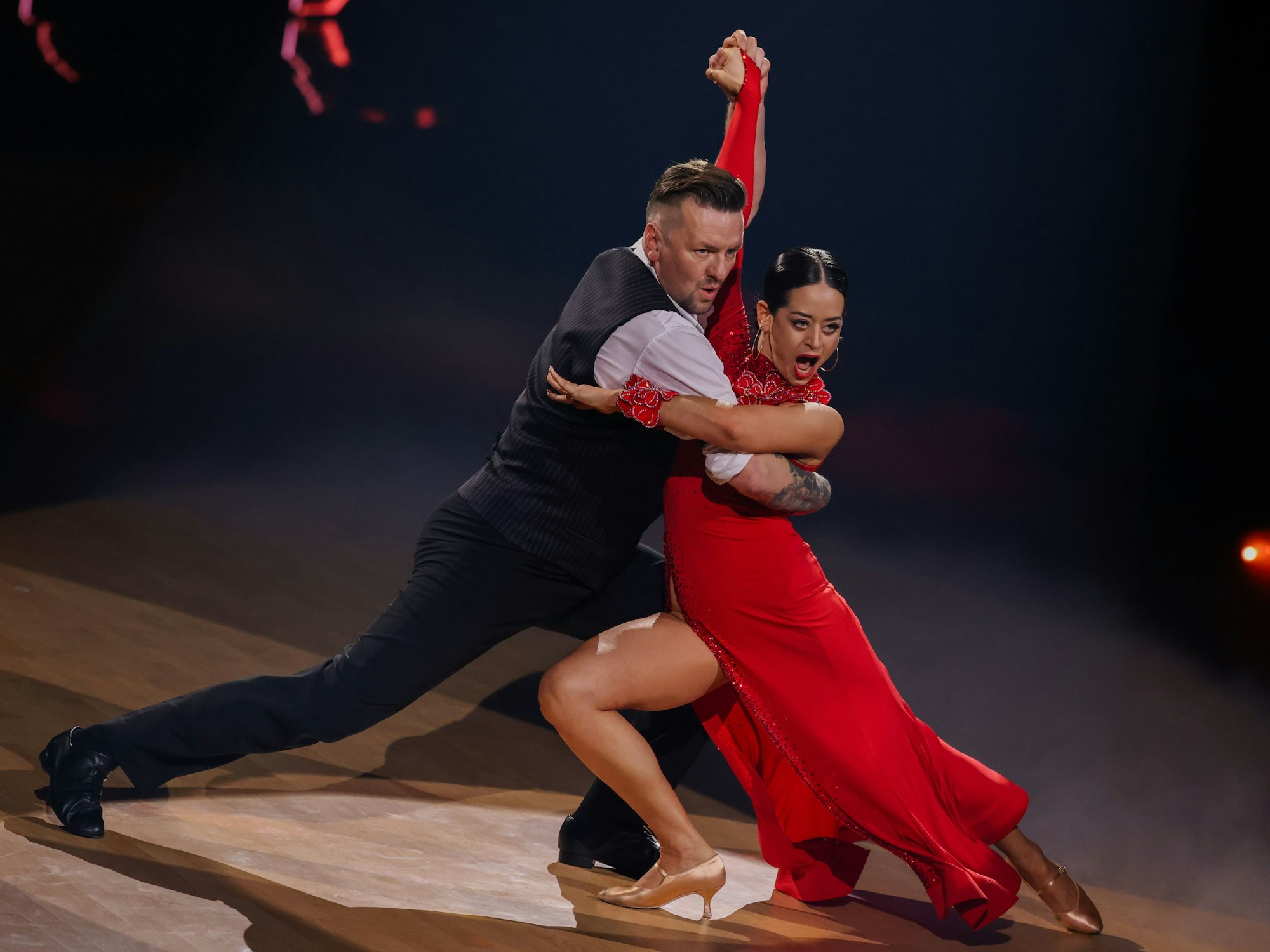 Ein Tanzpaar auf sdem Parkett. Die Frau trägt ein knallrotes Kleid, der Mann weißes Hemd mit Weste.