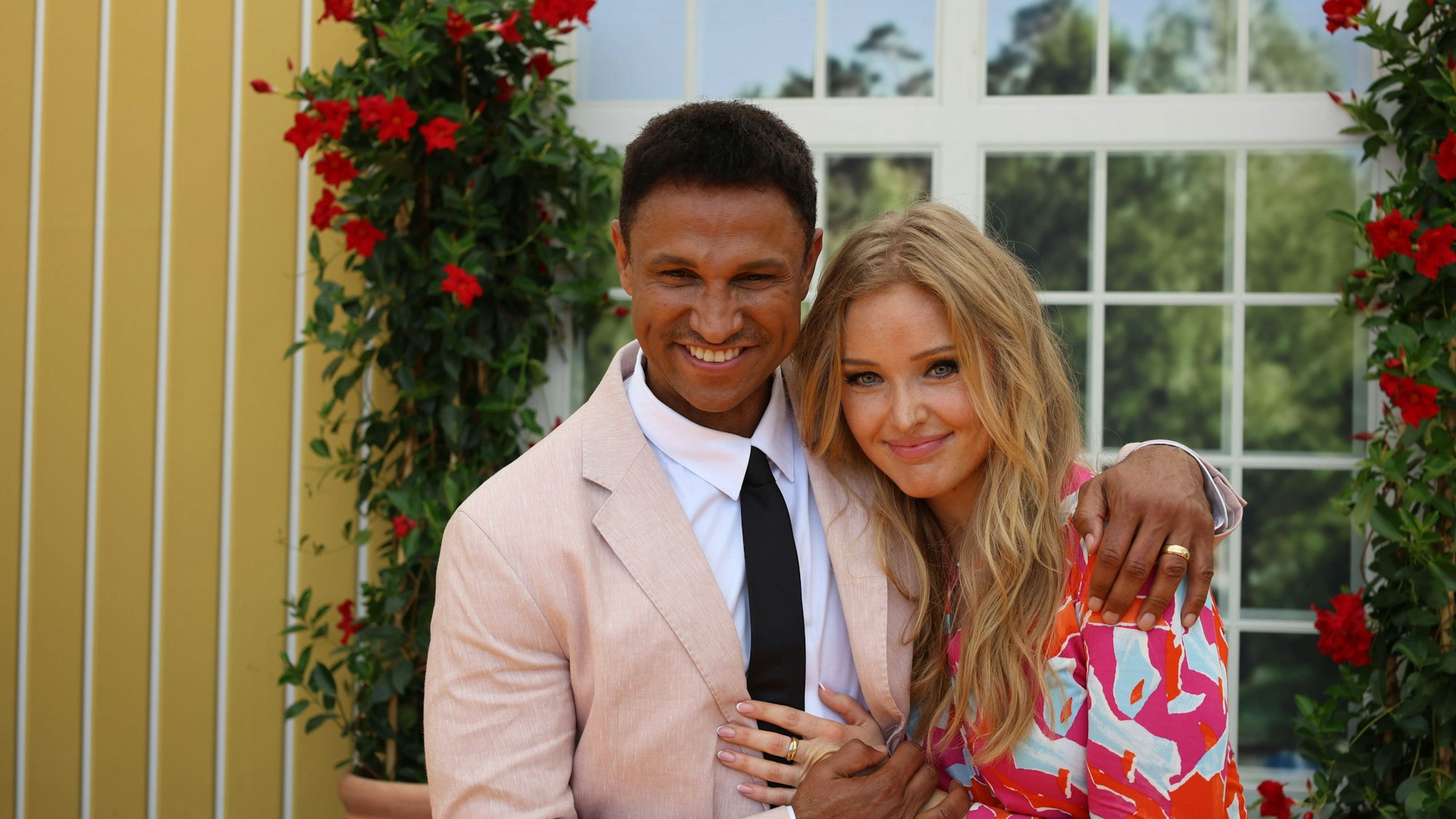 Patrice Aminati und Daniel Aminati lächeln glücklich, sich umarmend, vor Blüten und Fenster