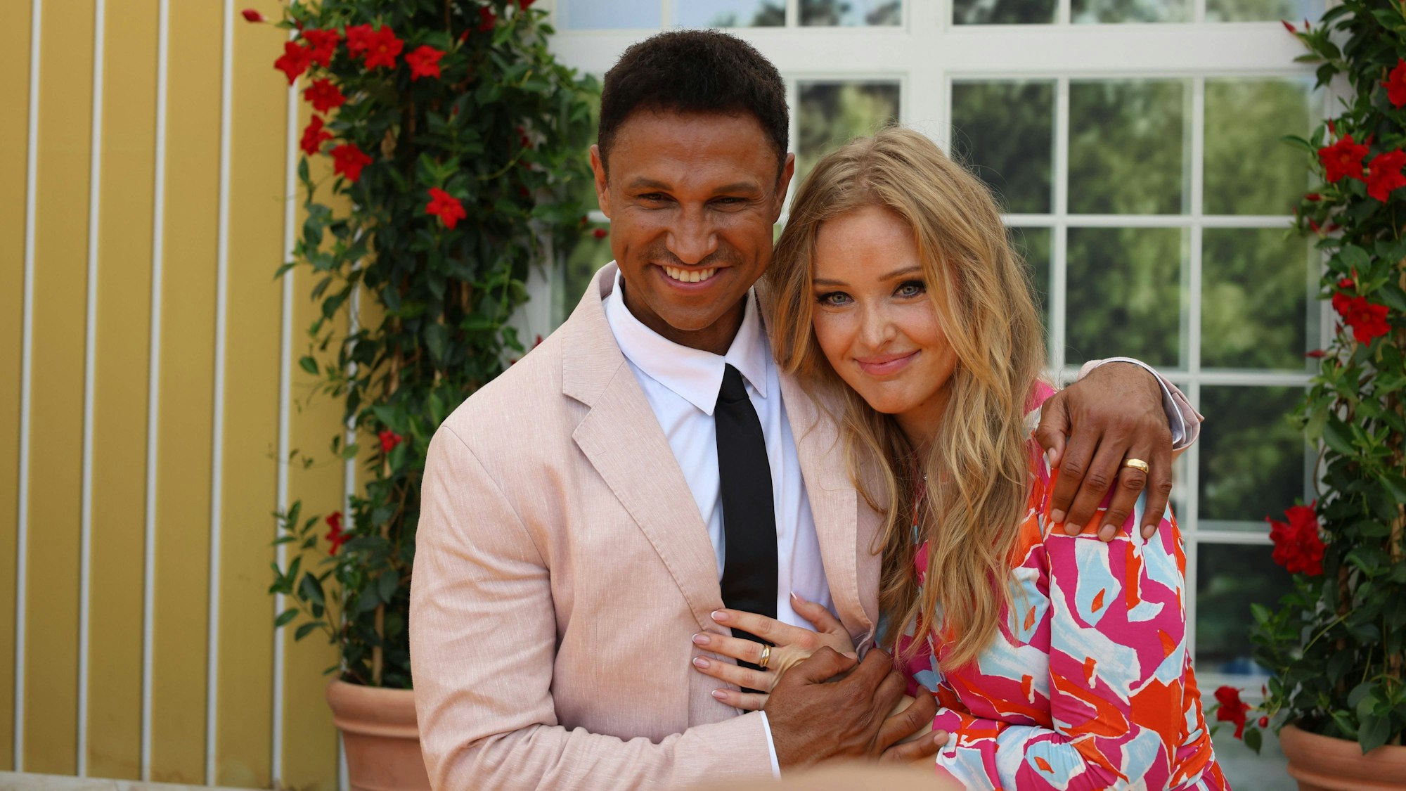 Patrice Aminati und Mann lächeln glücklich, sich umarmend, vor Blüten und Fenster