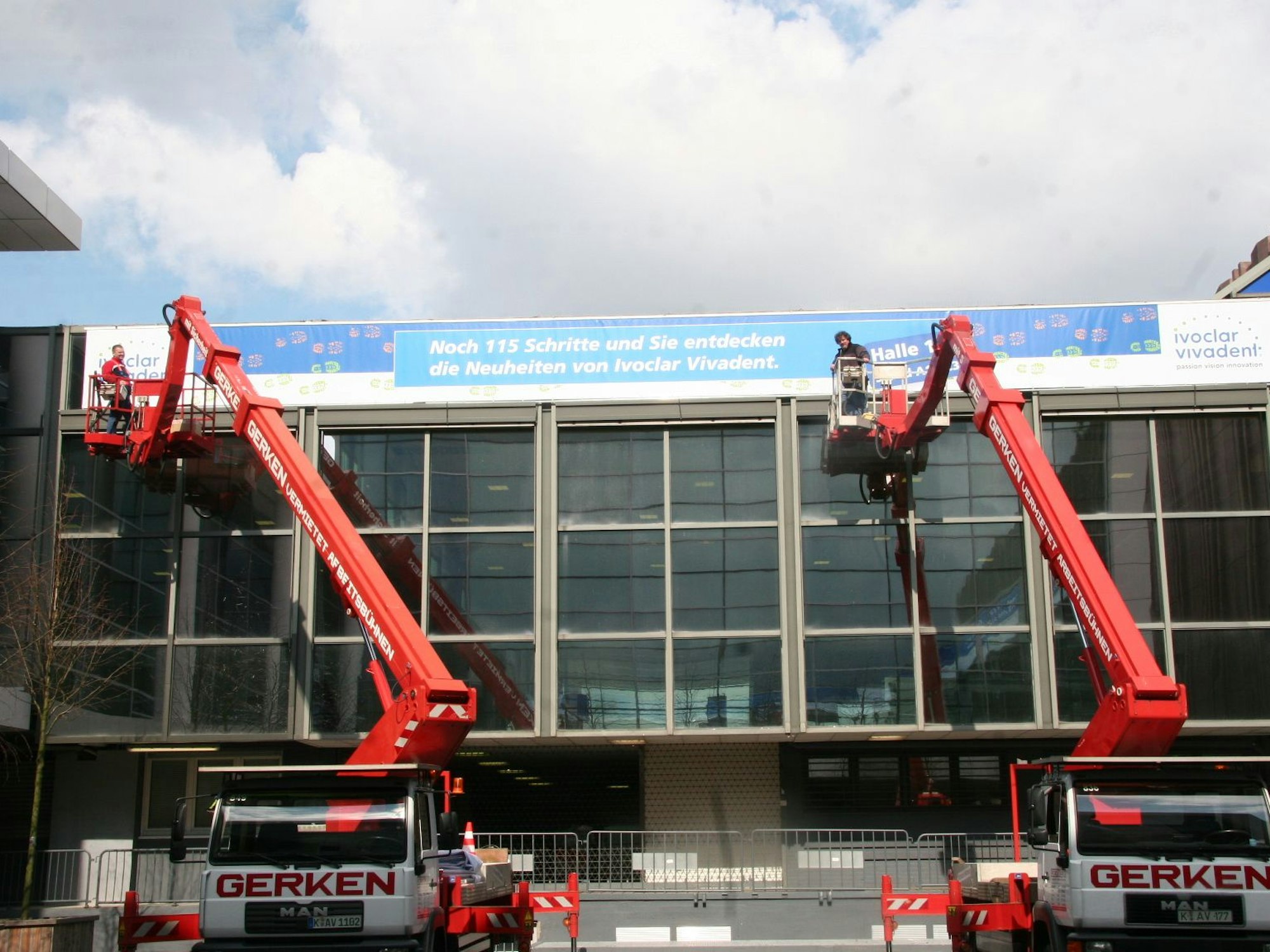 Zwei Arbeitsbühnen stehen ausgefahren außen an einer Halle, darauf jeweils ein Mann.