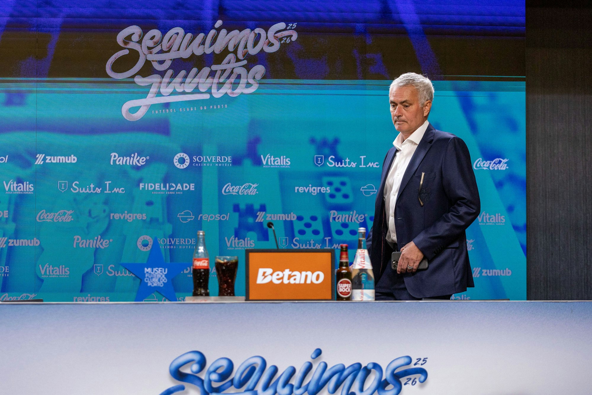 José Mourinho geht an Sponsorentafel vorbei mit Getränken und Schriftzug „Seguimos Juntos“.
