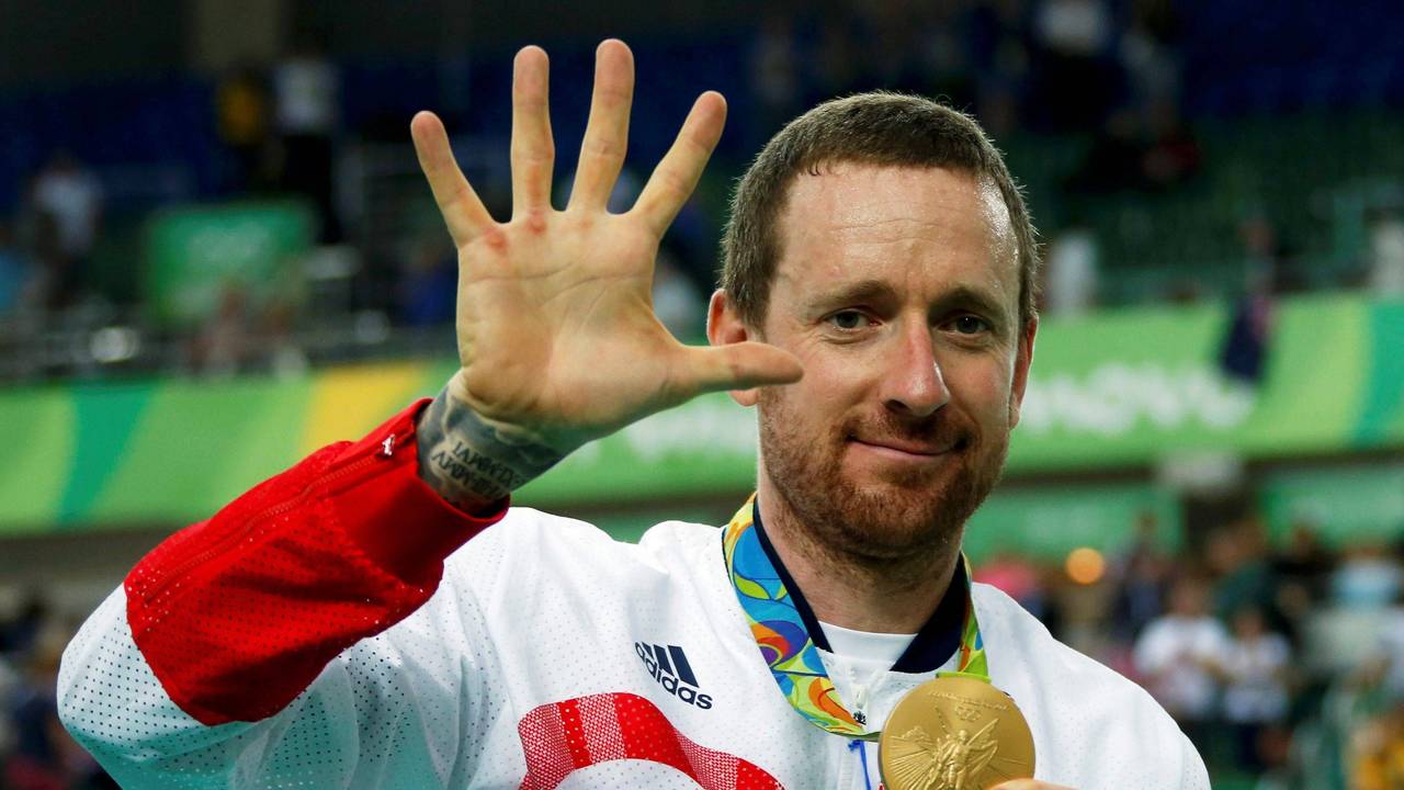 Bradley Wiggins zeigt seine olympische Goldmedaille in die Kamera und zeigt mit seiner rechten Hand die Zahl fünf.