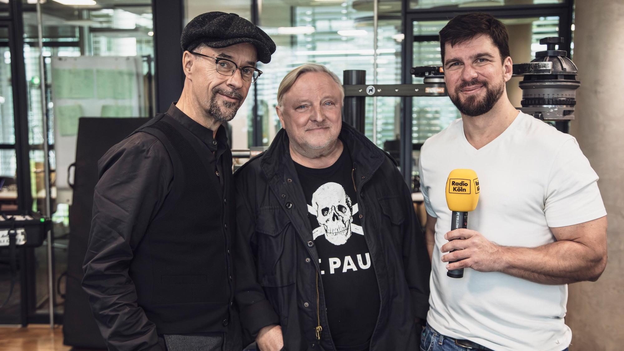 Zwei Schauspieler stehen neben einem Radiomoderator.