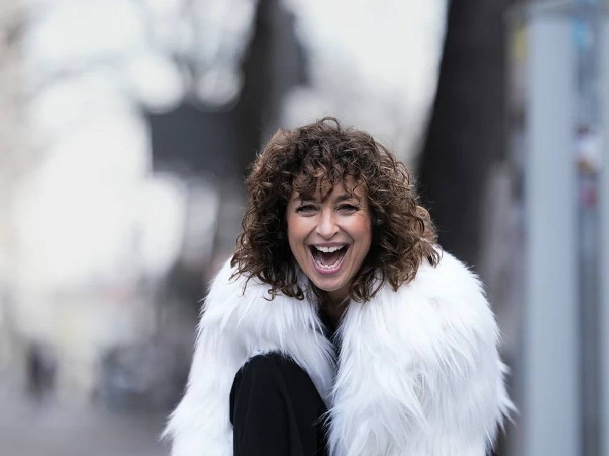 Eine fröhliche Frau mit dunklen Locken und weißer Zotteljacke schaut dynamisch lachend in die Kamera.