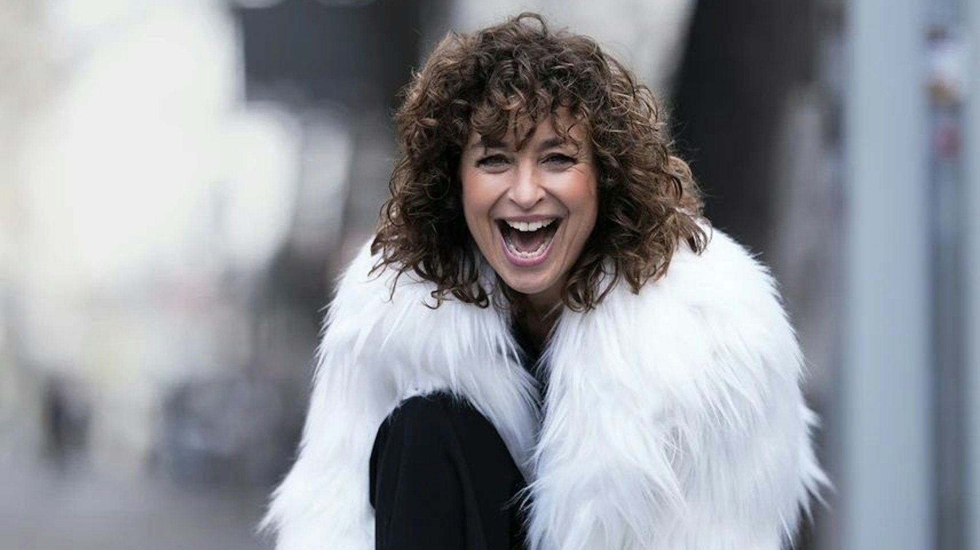 Eine fröhliche Frau mit dunklen Locken und weißer Zotteljacke schaut dynamisch lachend in die Kamera.