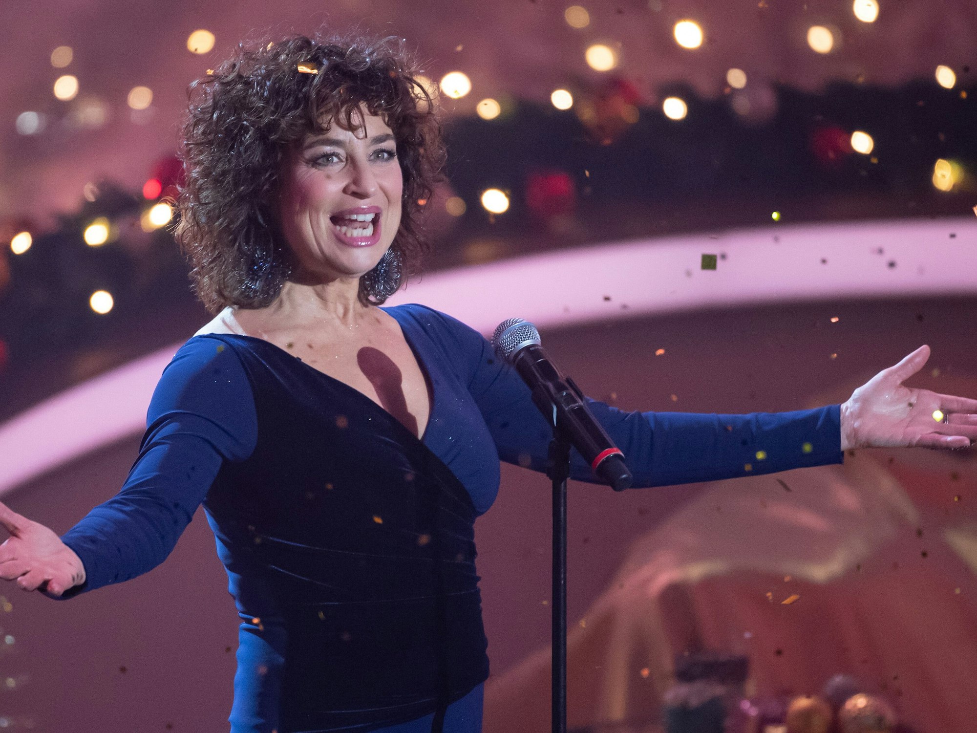 Eine Sängerin im dunkelblauen Kleid mit dunkeln Locken singt auf einer Bühne vor einem Mikrofon.