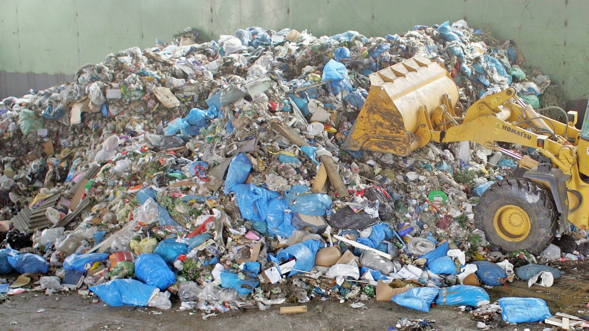 Ein gelber Bagger fährt in den Müll, der sich in einer Hausmülldeponie stapelt.