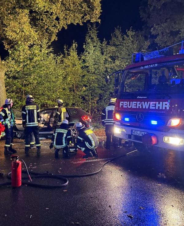 Einsatzkräfte von Feuerwehr und Rettungsdienst stehen an einem Auto, das vor einen Baum gefahren ist.