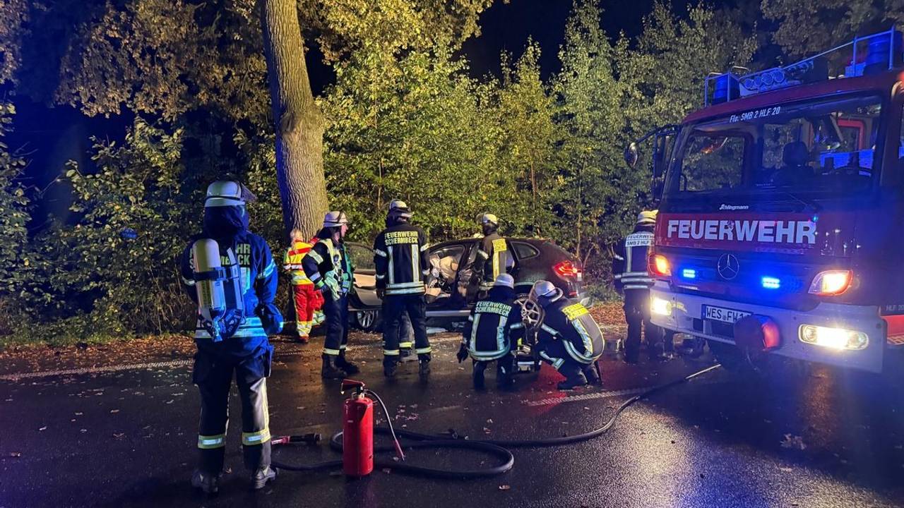 Einsatzkräfte von Feuerwehr und Rettungsdienst stehen an einem Auto, das vor einen Baum gefahren ist.