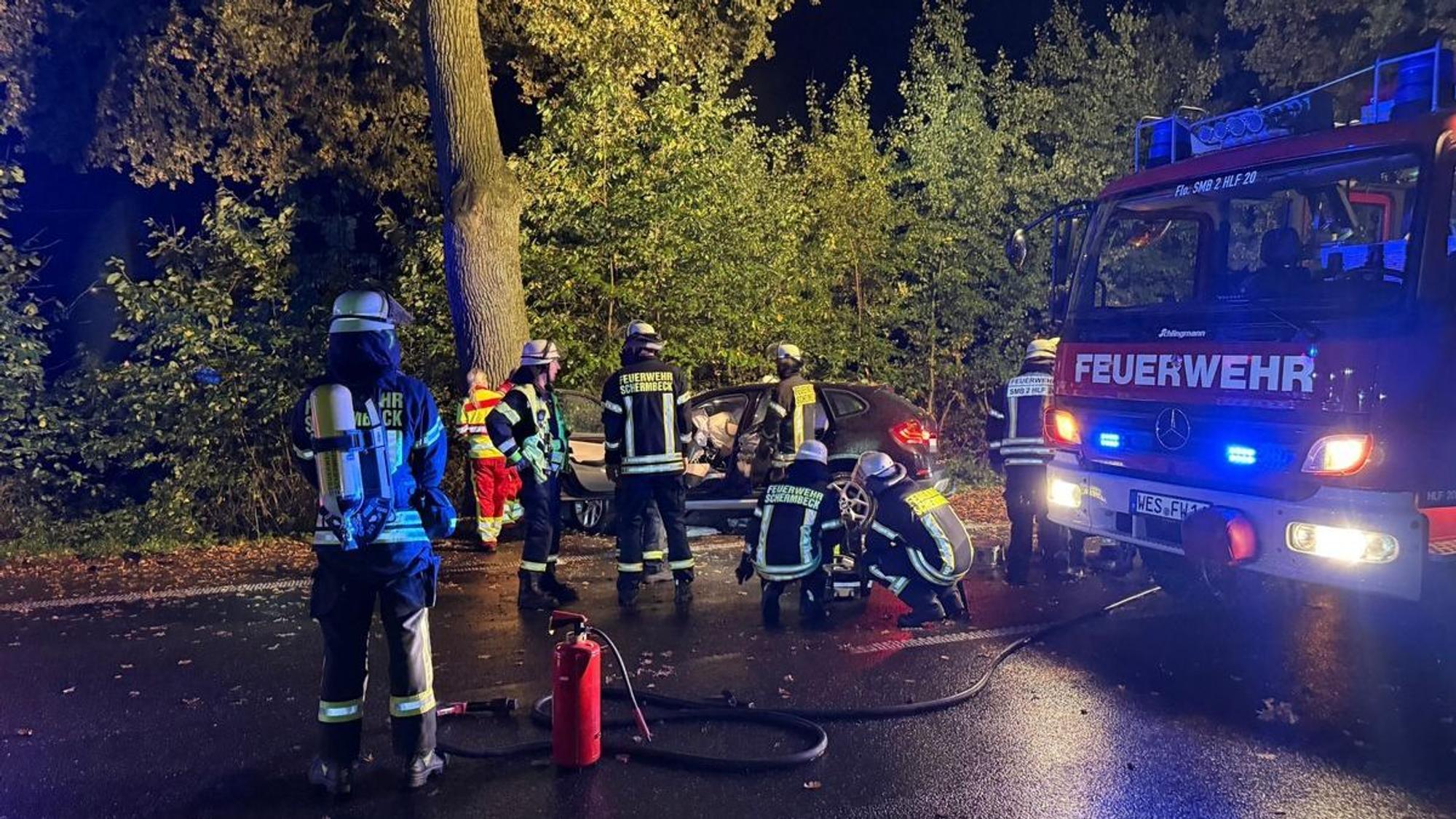Einsatzkräfte von Feuerwehr und Rettungsdienst stehen an einem Auto, das vor einen Baum gefahren ist.