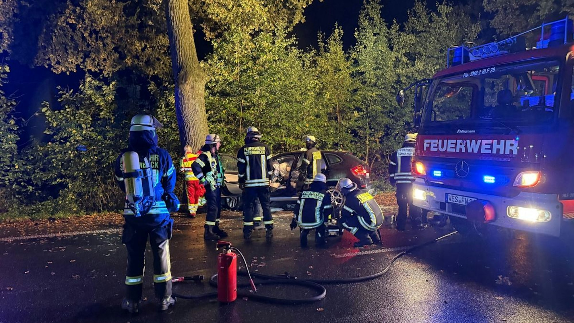 Einsatzkräfte von Feuerwehr und Rettungsdienst stehen an einem Auto, das vor einen Baum gefahren ist.