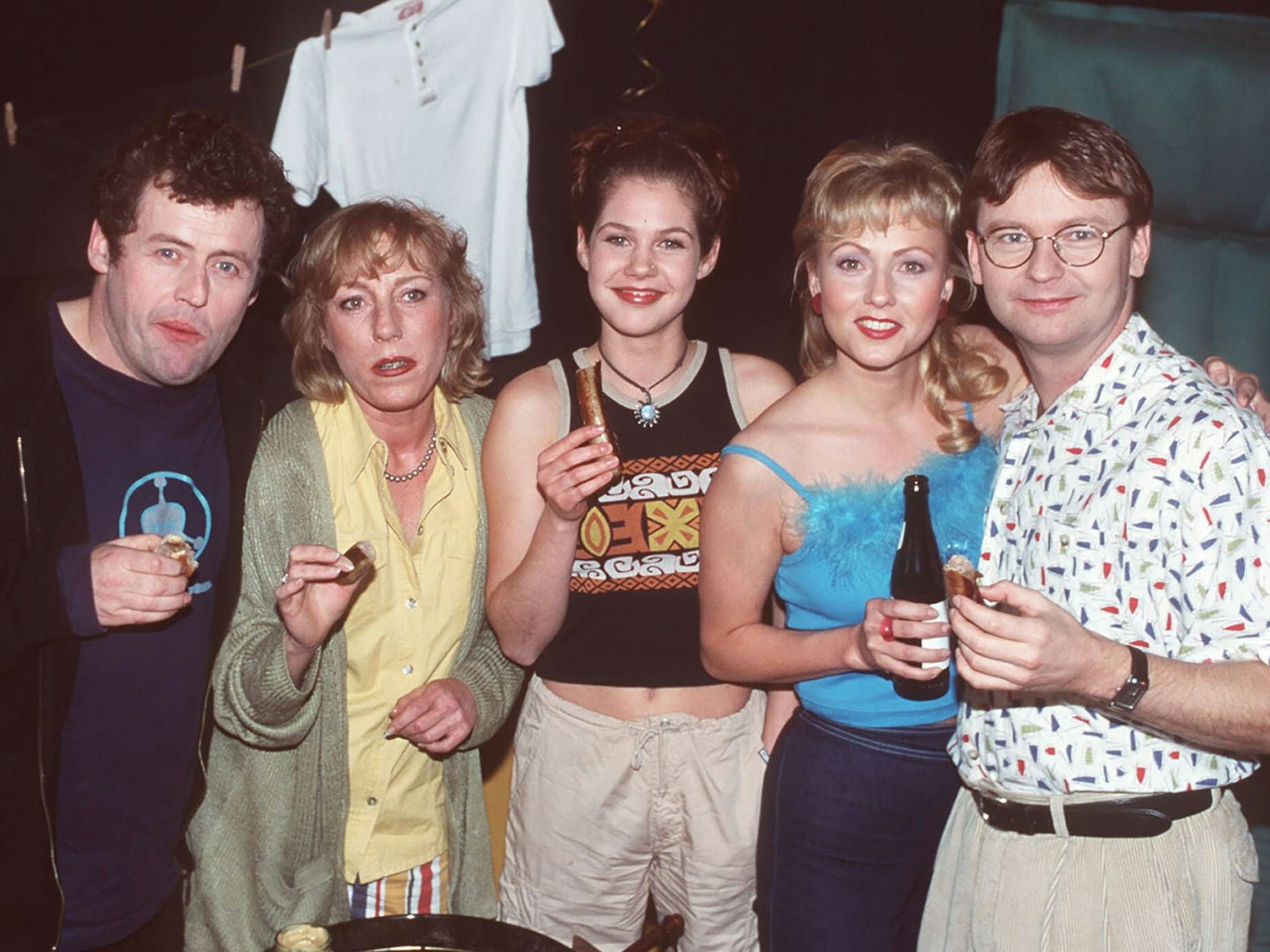 Füng Personen in 90er-Jahre-Kleidung halten Bier und Bratwurst in den Händen und schauen teils kritisch, teils belustigt, in die Kamera.