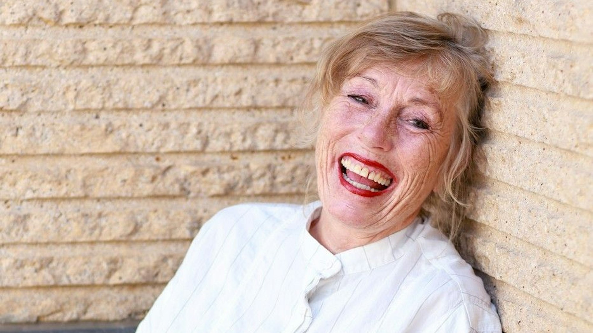 Eine Frau mittleren Alters mit knallroten Lippen lehnt an einer Steinmauer und lacht.