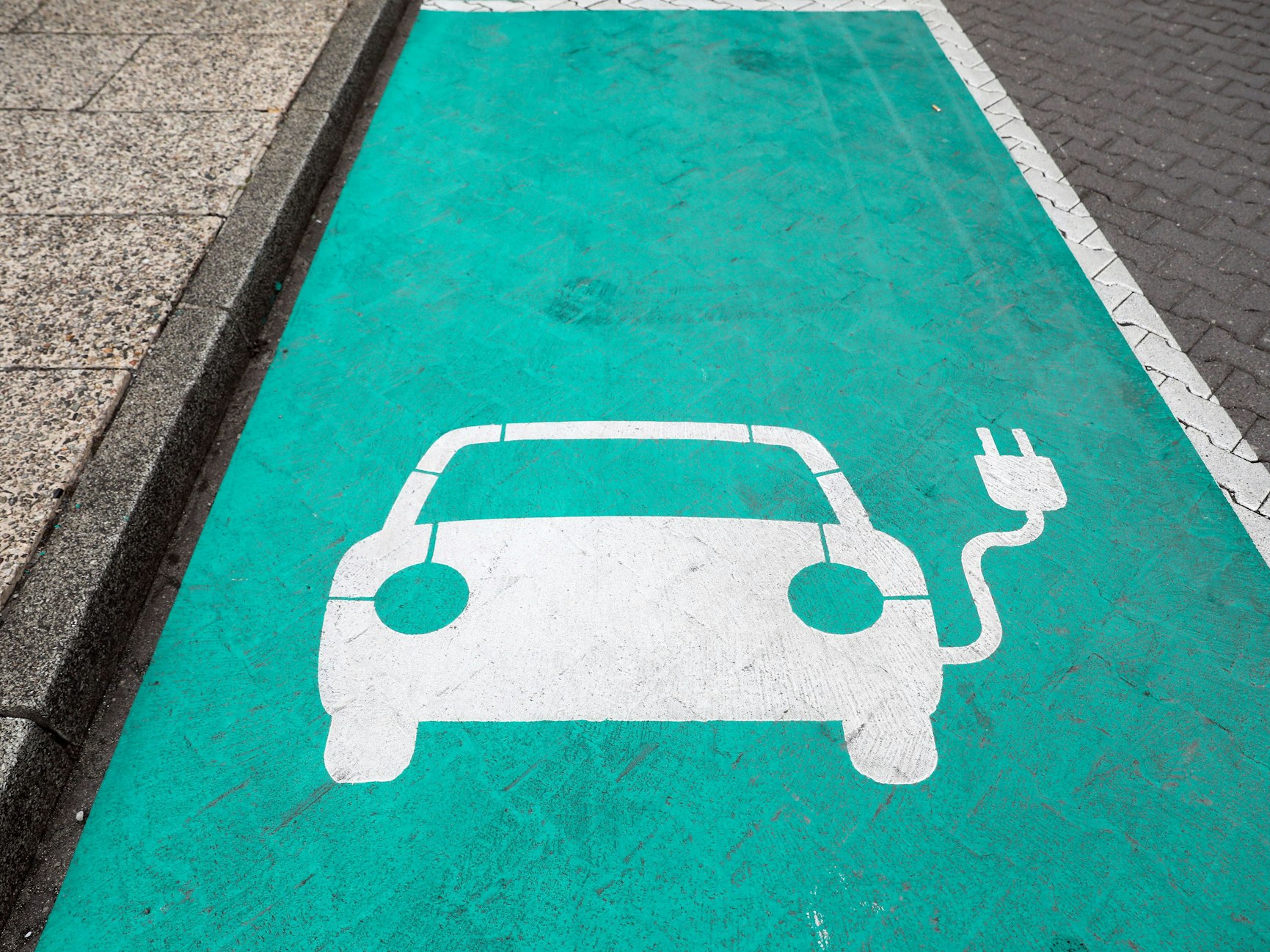 Ein stilisiertes Auto auf einem grünen Grund markiert einen Parkplatz mit Ladesäule für Elektrofahrzeuge.