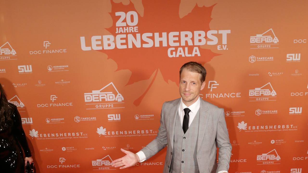 Ein Mann im Anzug steht vor einer Wand mit der Aufschrift Lebensherbst.