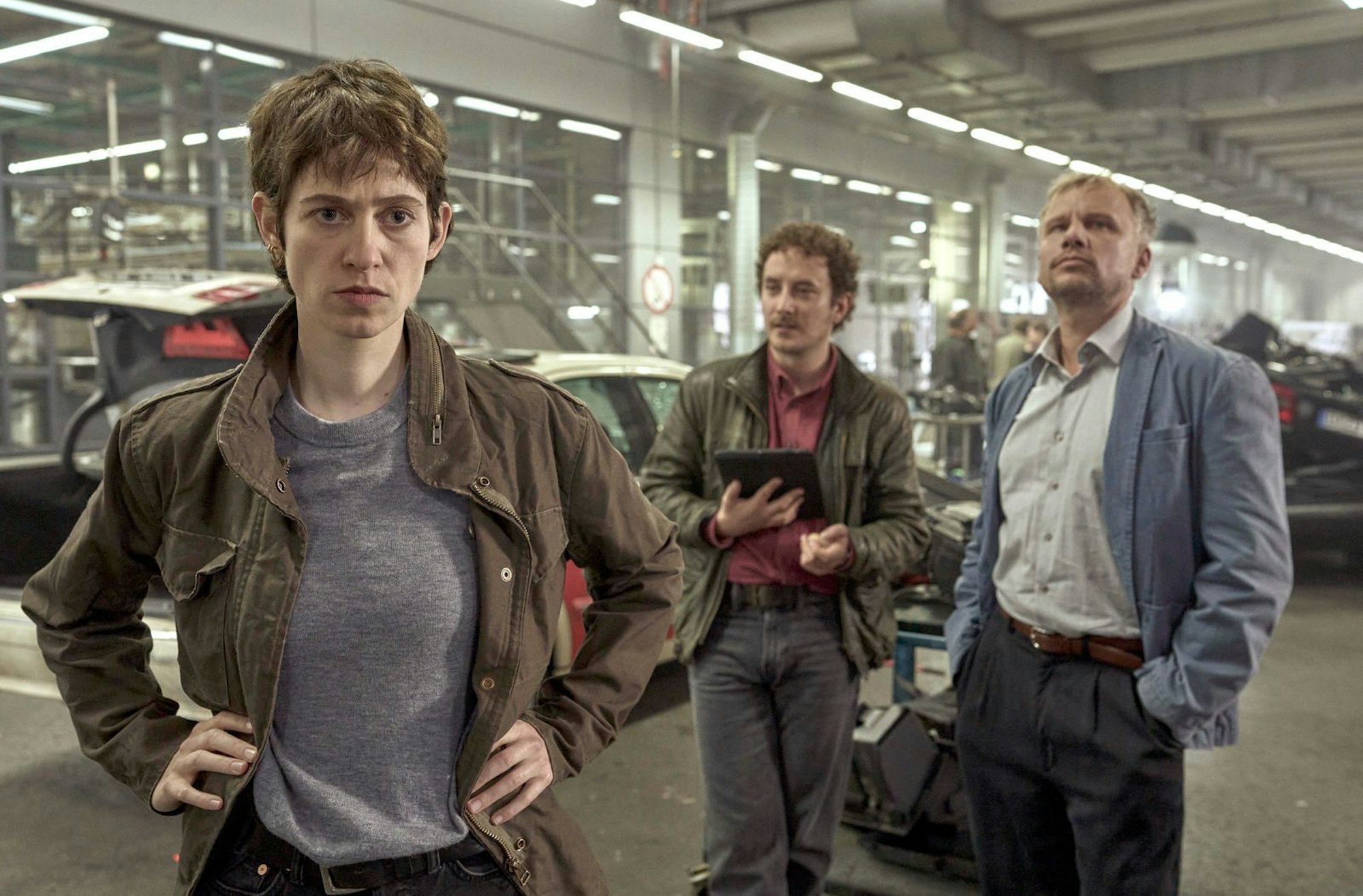Die Ermittler Anne Goldmundt (Lia von Blarer), Hänno Gudjons (David Hugo Schmidt) und Jan Auschra (Robert Stadlober, rechts) arbeiten in einer großen Halle, in der sämtliche Autowracks zu Untersuchungszwecken „aufgebahrt“ wurden. (Bild: WDR/Windlight Pictures/Satel Film/Frank Dicks)