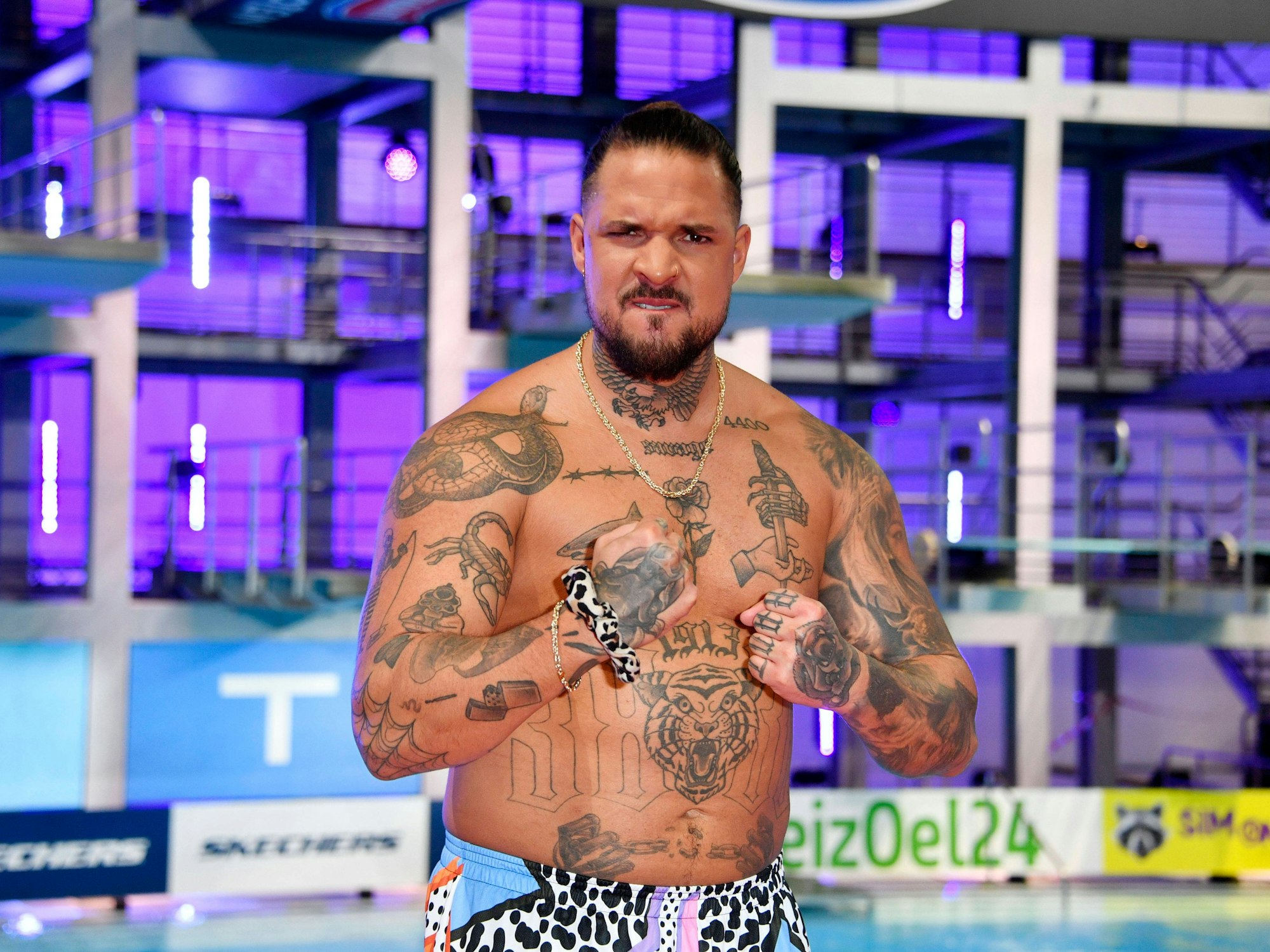 Zico Banach schaut mit angespanntem Gesicht in die Kamera, trägt nur eine Badehose und präsentiert seine Muskeln und Tattoos.