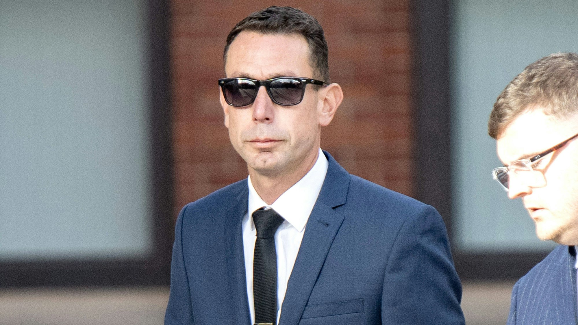 „David Coote“ im Anzug und Sonnenbrille blickt ernst in die Kamera