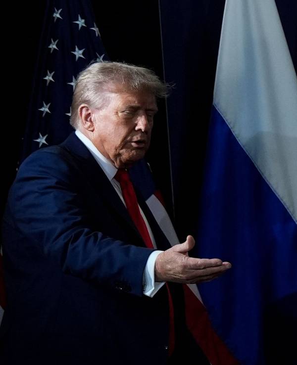 Donald Trump hält seine Hand einladend nach vorne, Putin daneben ist fast verdeckt, nur seine obere Kopfhälfte ragt über einem Plakat hervor.