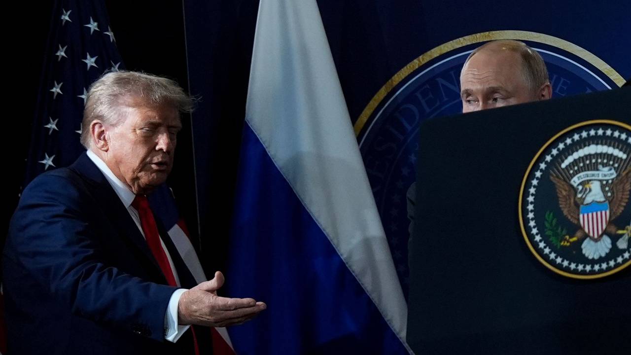 Donald Trump hält seine Hand einladend nach vorne, Putin daneben ist fast verdeckt, nur seine obere Kopfhälfte ragt über einem Plakat hervor.