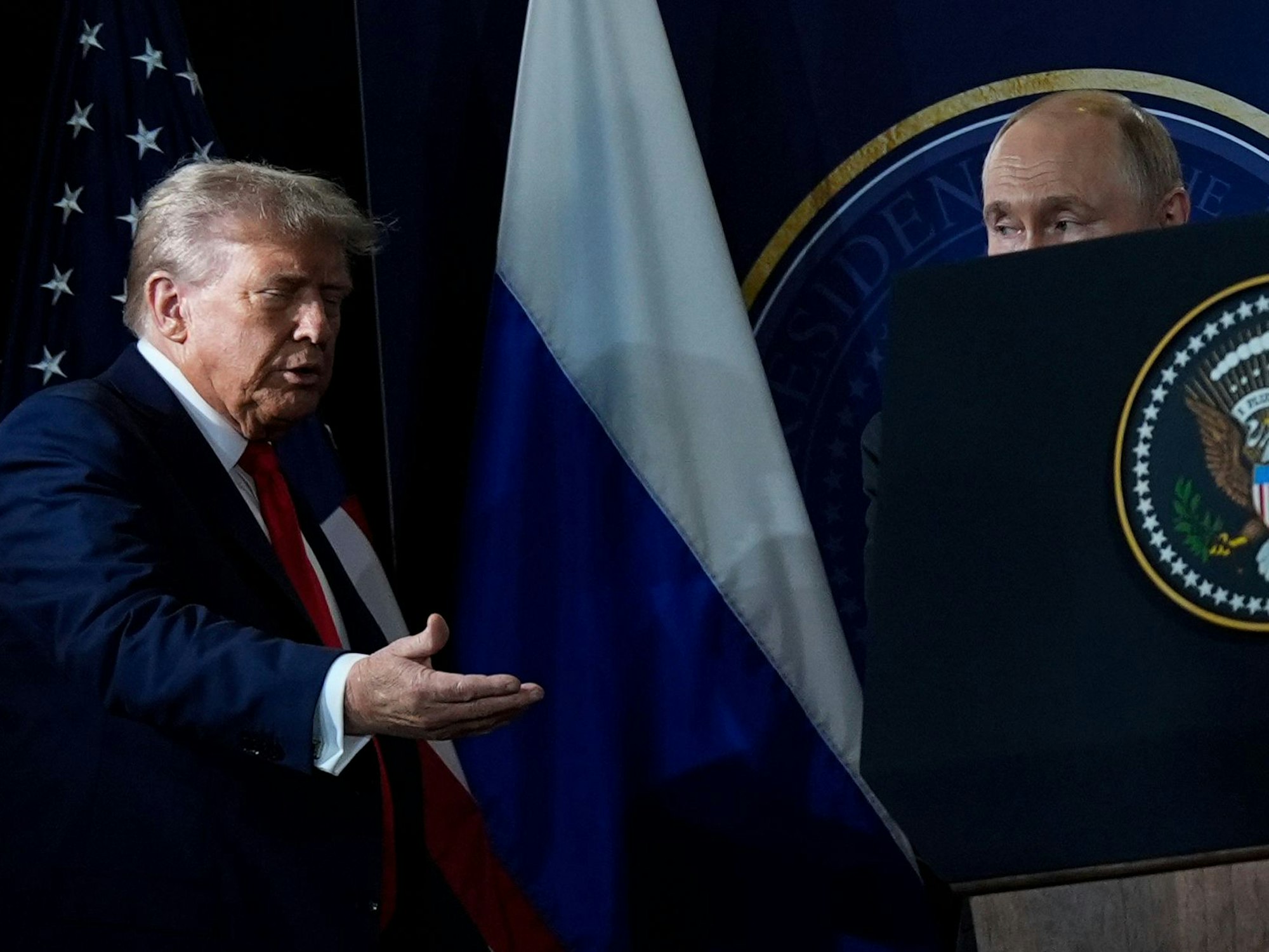 Donald Trump hält seine Hand einladend nach vorne, Putin daneben ist fast verdeckt, nur seine obere Kopfhälfte ragt über einem Plakat hervor.